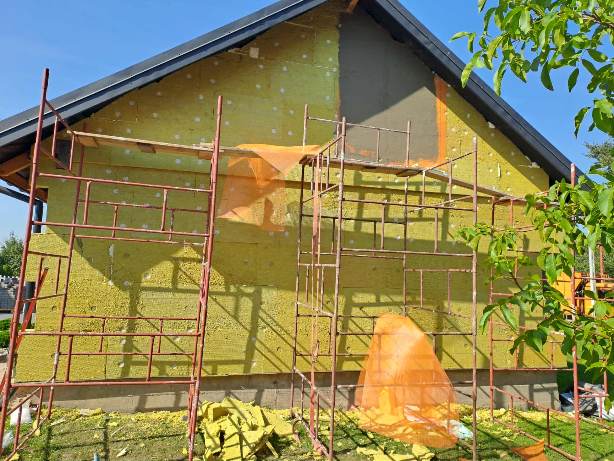 Dom w trakcie ocieplania: żółta wełna mineralna na ścianach, częściowo pokryta szarą zaprawą. Widoczne rusztowania i fragmenty wełny na trawie. Słoneczny dzień.