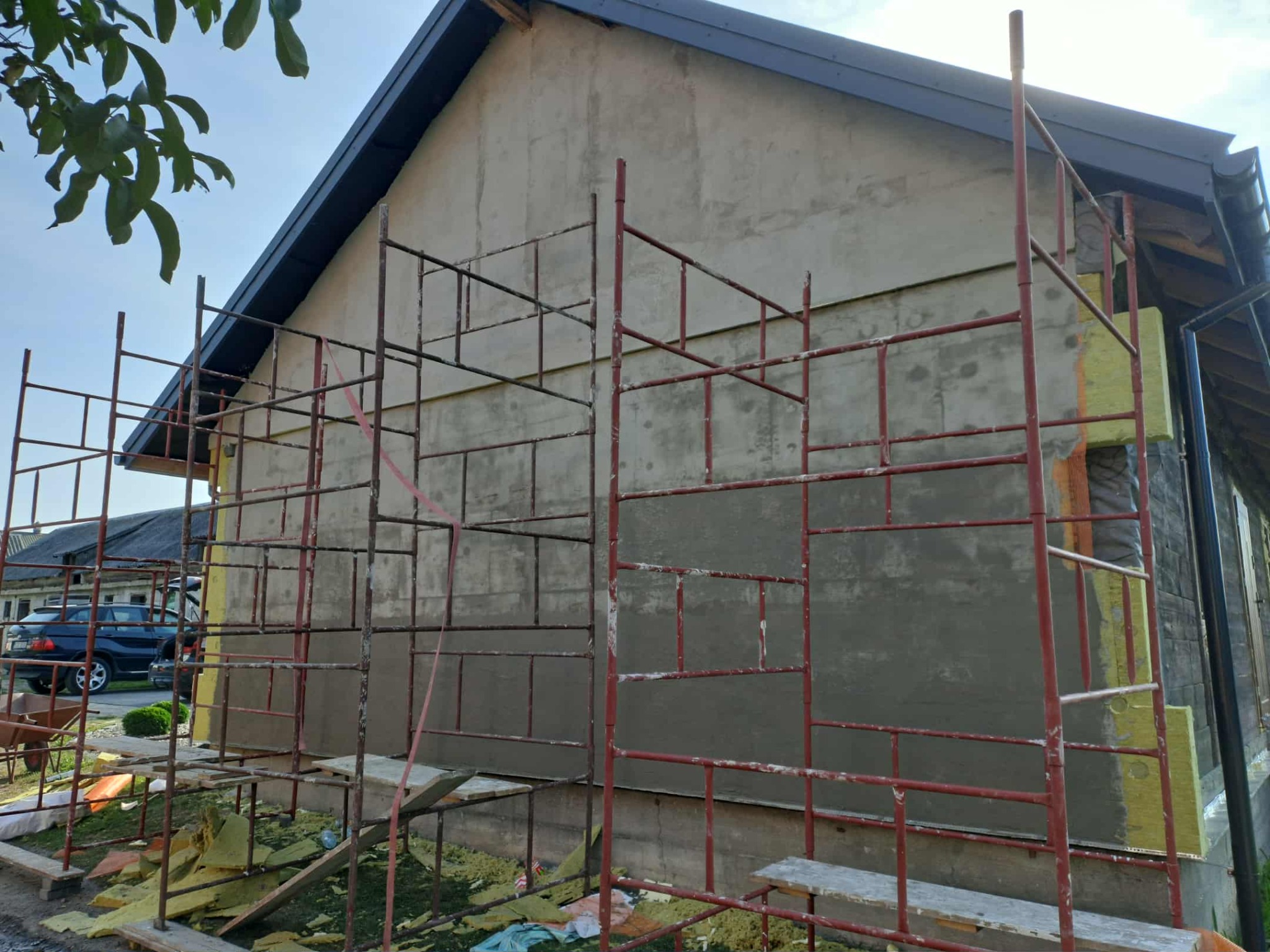 Budynek w trakcie prac ociepleniowych, widoczne rusztowania i fragment ściany z izolacją. Częściowo nałożony tynk. Elementy ocieplenia leżą na ziemi.