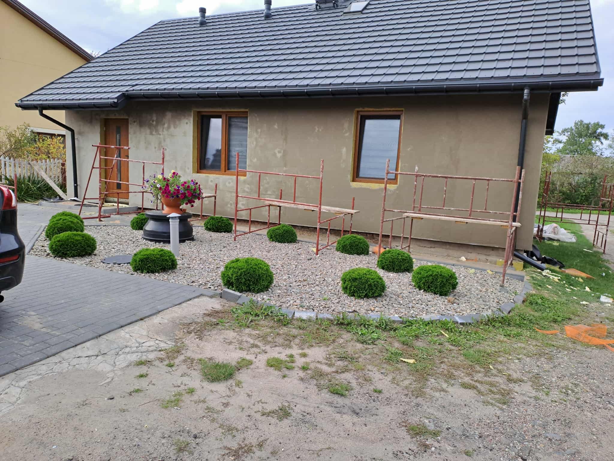 Dom jednorodzinny w trakcie prac elewacyjnych, widoczne rusztowania, ocieplenie wokół okien i ozdobne krzewy w otoczeniu budynku.