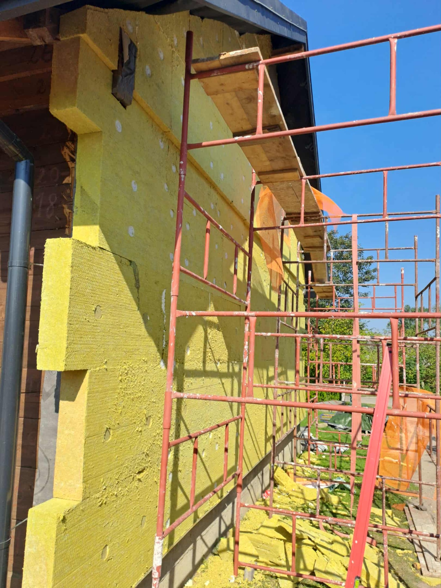 Ocieplanie ściany budynku żółtą wełną mineralną, widoczne rusztowania i fragmenty materiału na ziemi. Prace w toku przy słonecznej pogodzie.