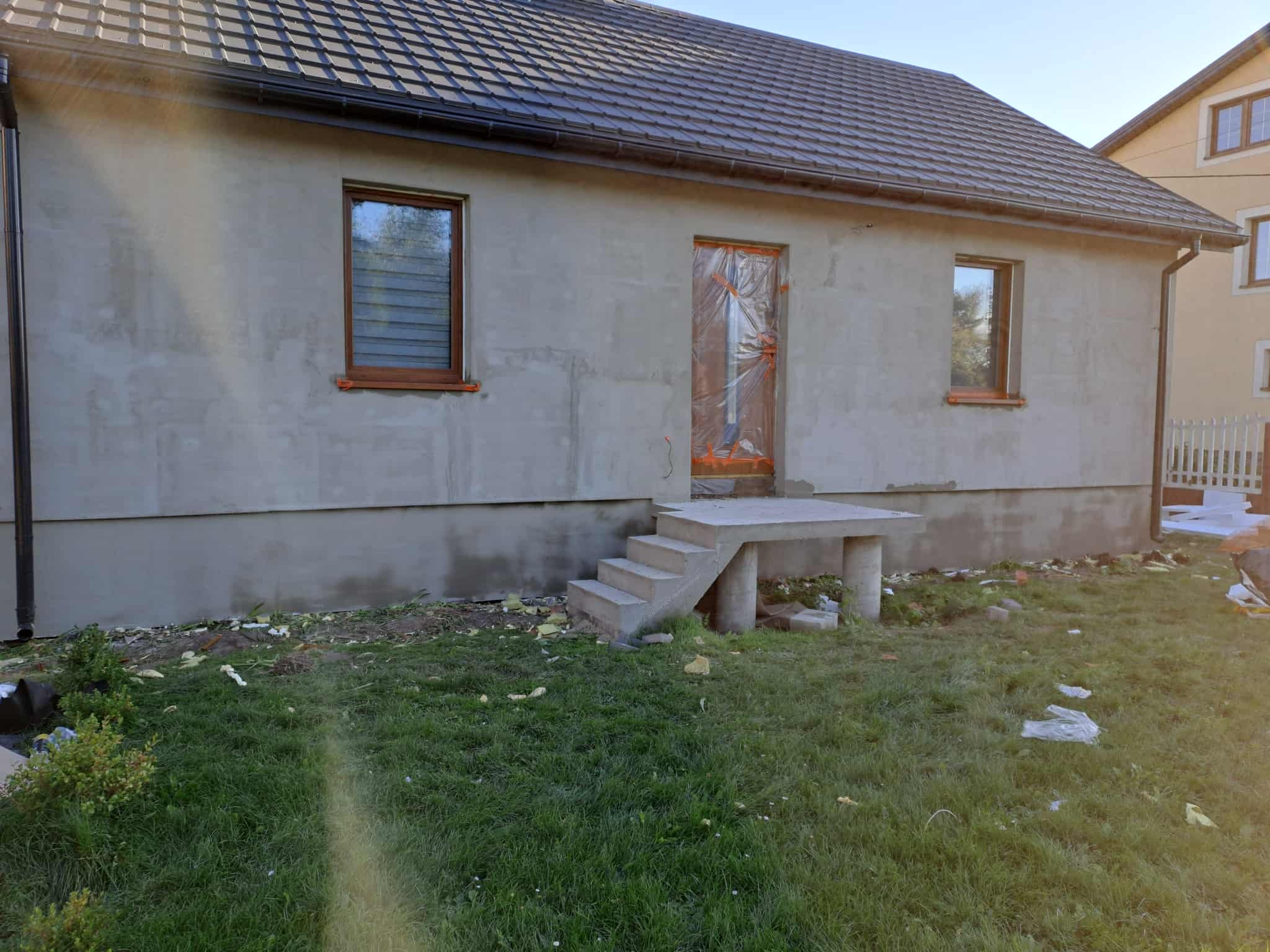 Szara elewacja domu jednorodzinnego w trakcie prac wykończeniowych, z oknami w drewnianych ramach i zabezpieczonym wejściem folią malarską.