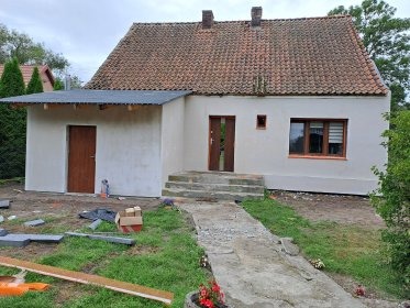 Odnowiony dom jednorodzinny z nową elewacją w jasnym odcieniu, widoczne schody wejściowe i ścieżka z kamienia. Częściowo widoczne rośliny w tle.