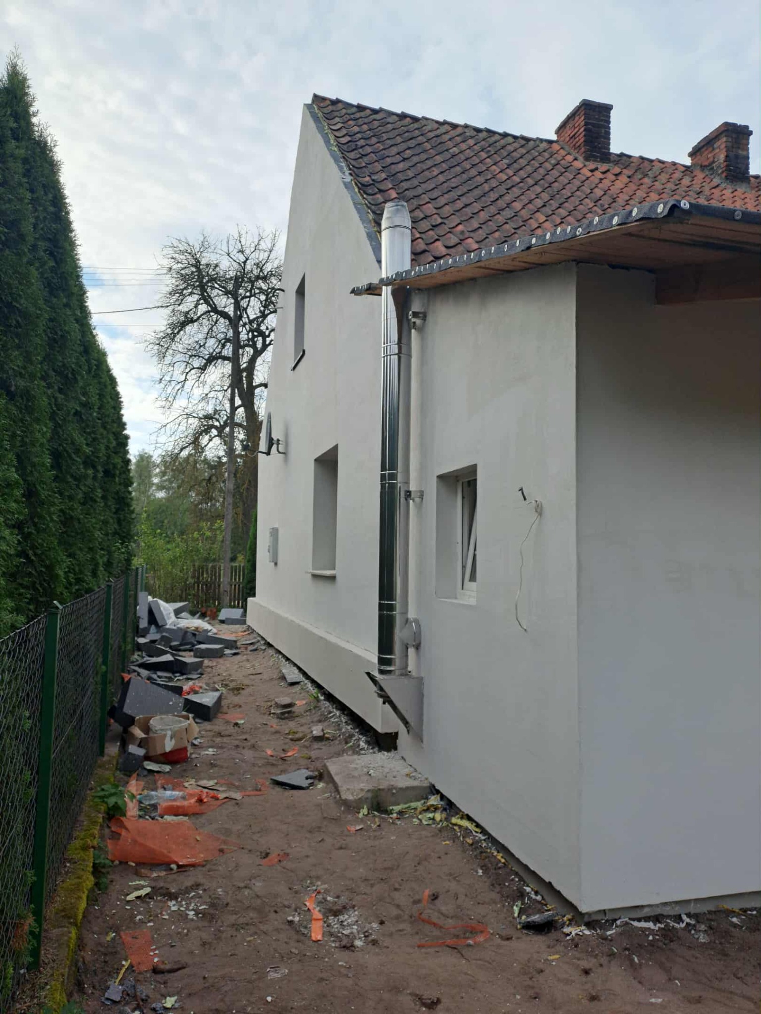 Boczny widok domu po malowaniu elewacji, z widocznym kominem ze stali nierdzewnej i fragmentami rozbiórki kostki brukowej wzdłuż ściany.