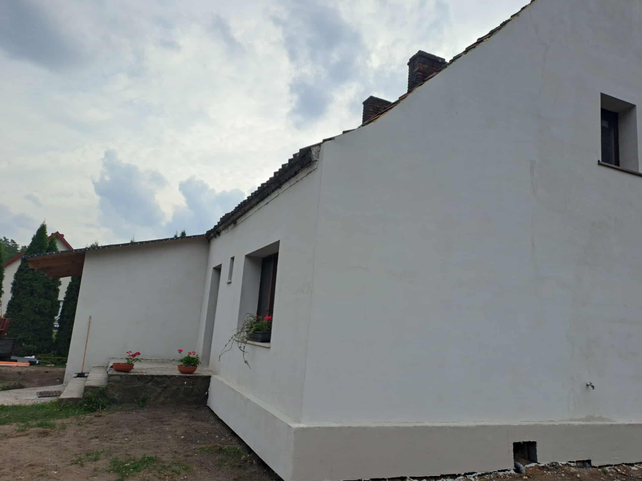 Biała elewacja domu jednorodzinnego po malowaniu, widok z boku, z kwiatami w doniczkach na schodach i pod oknem. Pochmurne niebo w tle.