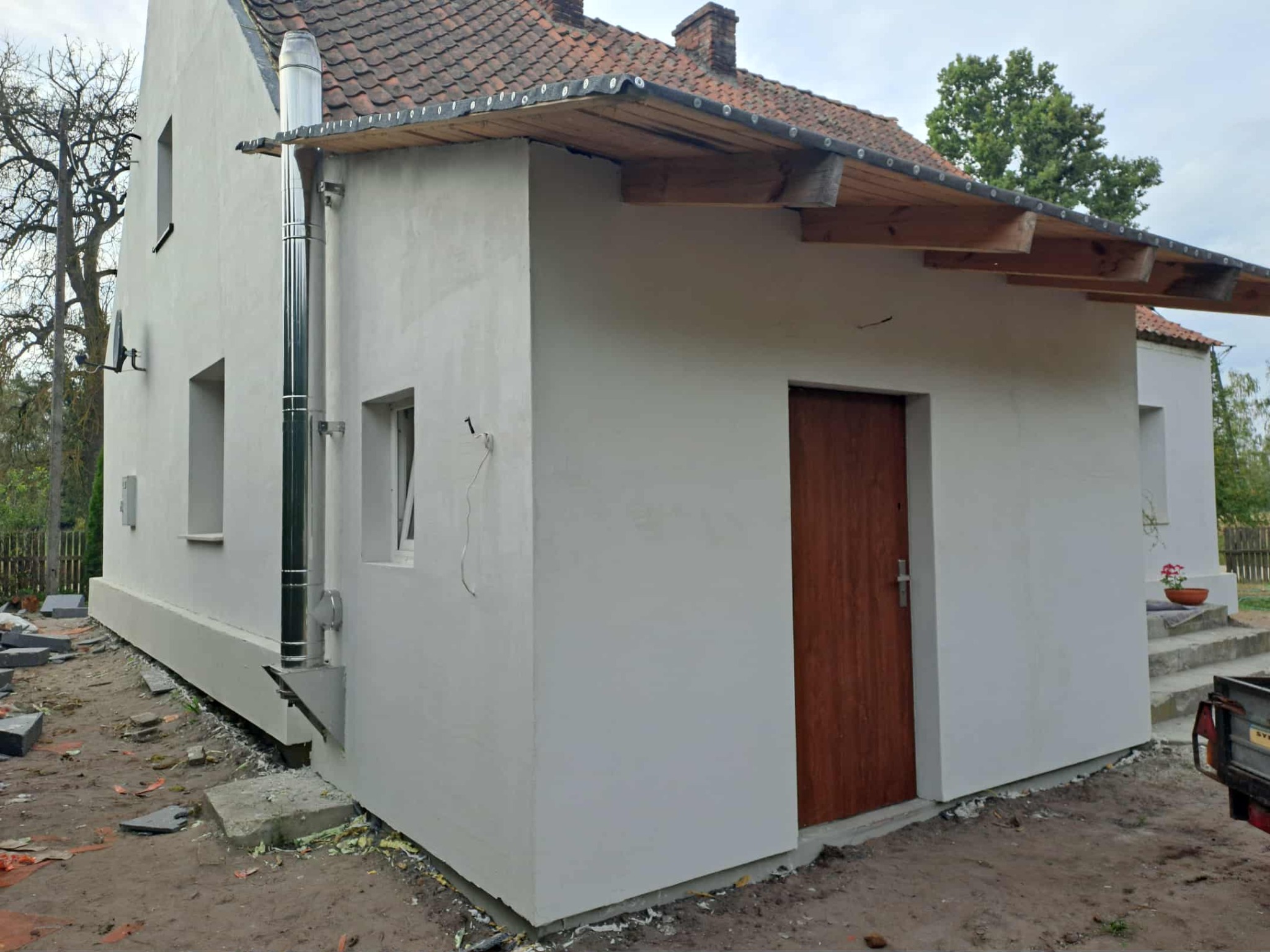 Elewacja budynku po remoncie, świeżo otynkowana na biało, z nowym kominem ze stali nierdzewnej i drewnianym zadaszeniem. Widoczne kostki brukowe do ułożenia.