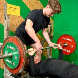 Gotowi Na Formę - Asystent asekuruje mężczyznę podczas wyciskania sztangi na ławce w siłowni. Obciążenia na sztandze w kolorach zielonym i czerwonym. Tabliczka informacyjna na ścianie.