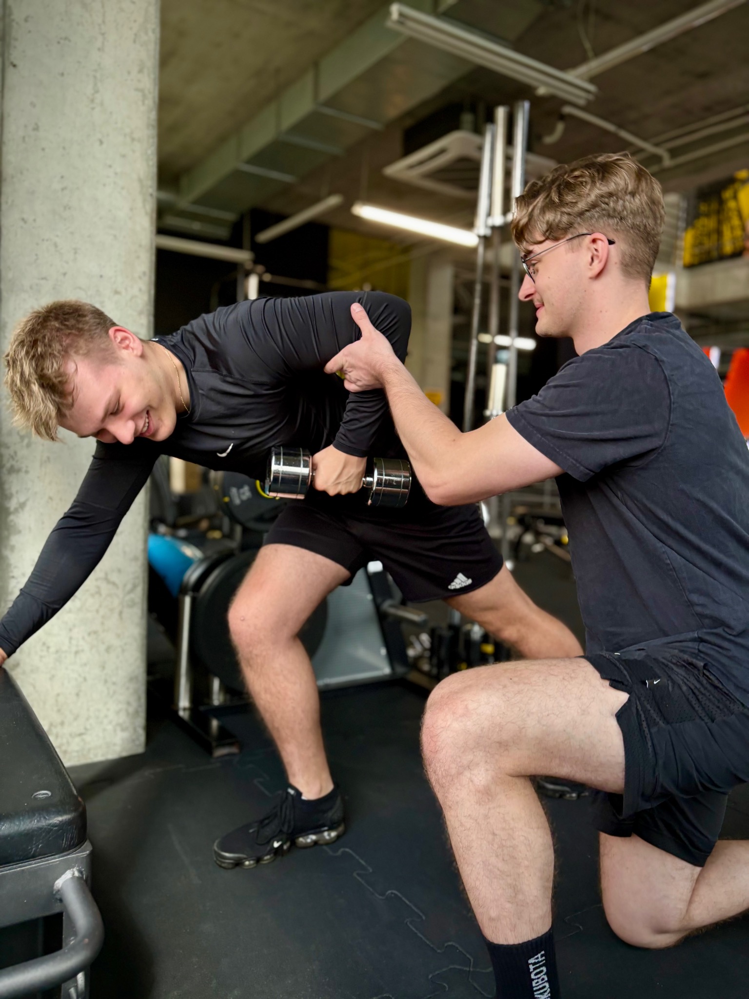 Trening personalny: mężczyzna ćwiczy podnoszenie hantli pod okiem trenera w czarnej koszulce, w siłowni z betonowym filarem w tle. Uśmiech na twarzy ćwiczącego.