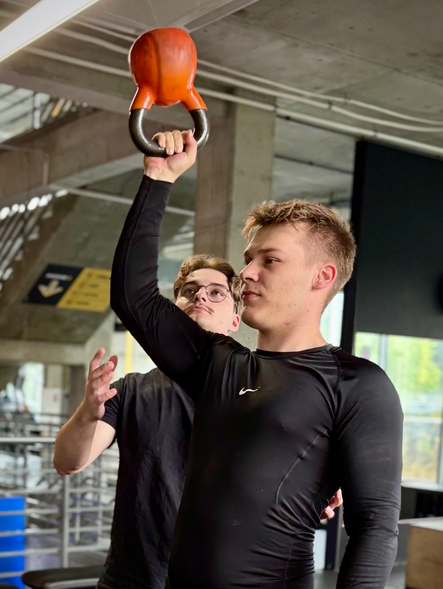 Młody mężczyzna w czarnej koszulce podnosi pomarańczowy kettlebell nad głowę, drugi mężczyzna obserwuje w tle, siłownia z betonowym sufitem.