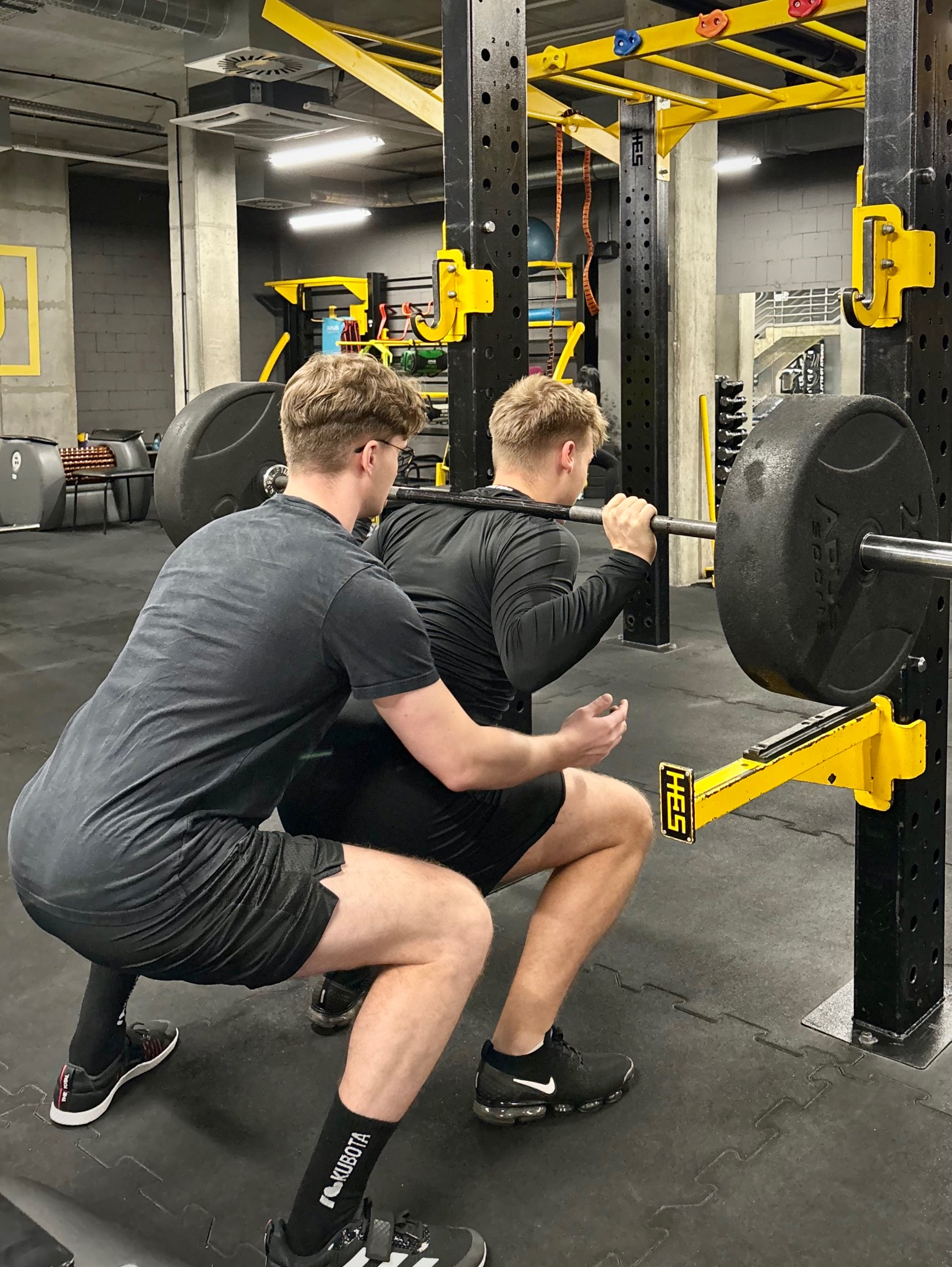 Mężczyzna w czarnej koszulce asekuruje drugiego podczas przysiadu ze sztangą w siłowni. Żółte elementy konstrukcji do ćwiczeń, gumowa podłoga.