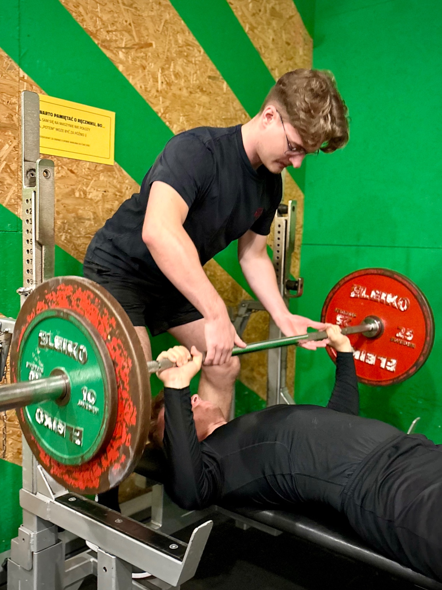Asystent asekuruje mężczyznę podczas wyciskania sztangi na ławce w siłowni. Obciążenia na sztandze w kolorach zielonym i czerwonym. Tabliczka informacyjna na ścianie.