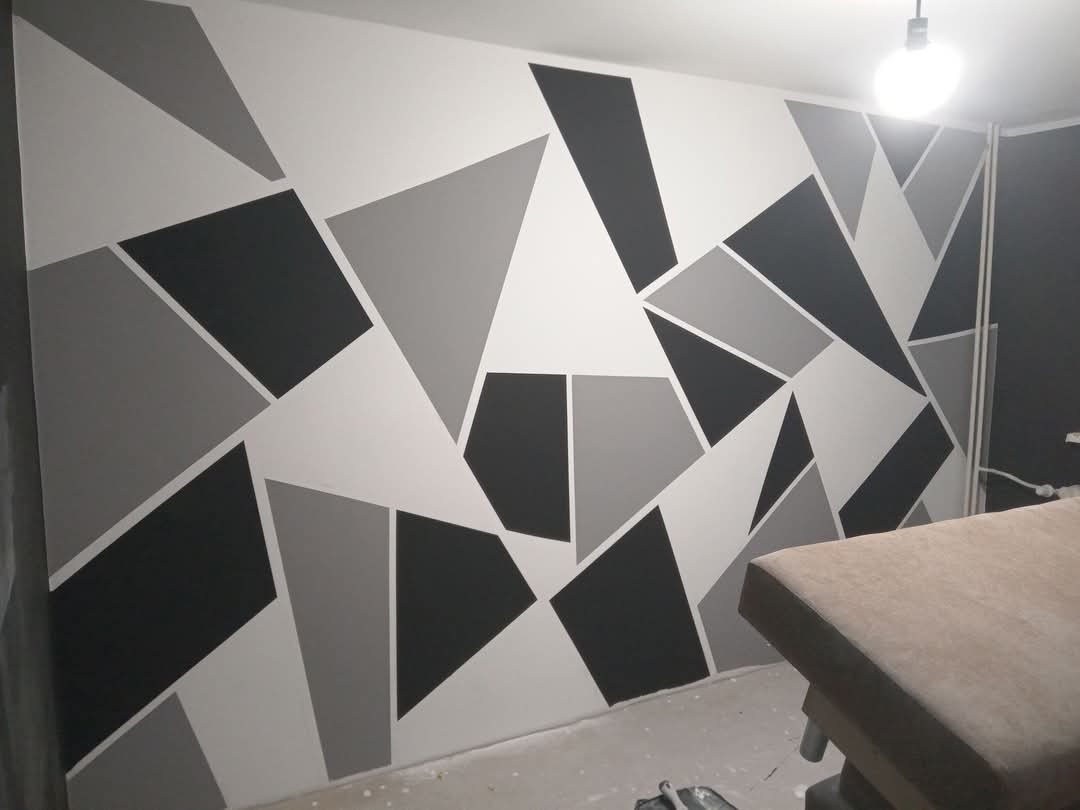 Ściana w pokoju pomalowana w geometryczne wzory w odcieniach szarości i bieli, oświetlona żarówką. Nowoczesny design wnętrza.