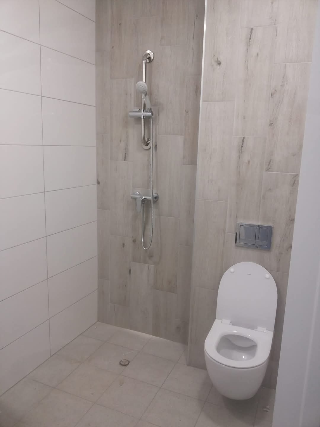 Nowoczesna łazienka po remoncie: białe płytki, imitacja drewna, toaleta wisząca i deszczownica. Minimalistyczny design, jasne wnętrze.