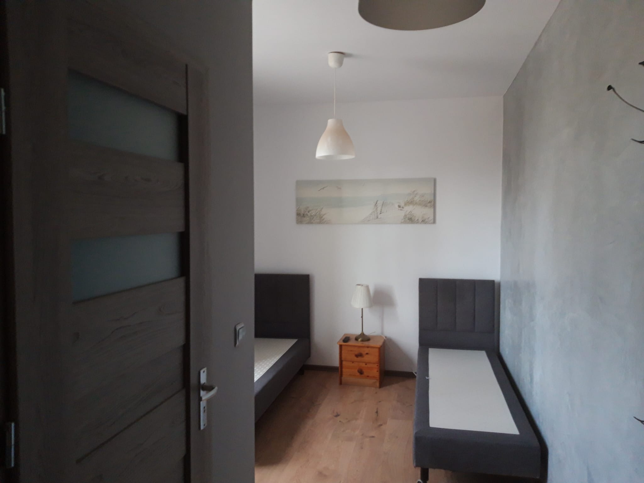 Jasne wnętrze sypialni z dwoma łóżkami, drewnianą podłogą i obrazem nad łóżkami. Widok przez drewniane drzwi z przeszkleniami. Szara ściana z dekoracyjnym wykończeniem.