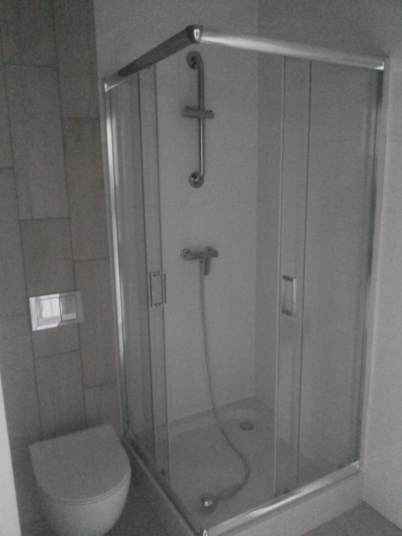 Nowoczesna kabina prysznicowa z chromowanymi profilami i szklanymi drzwiami, widoczna obok toaleta w łazience wykończonej szarą płytką. Minimalistyczny design.