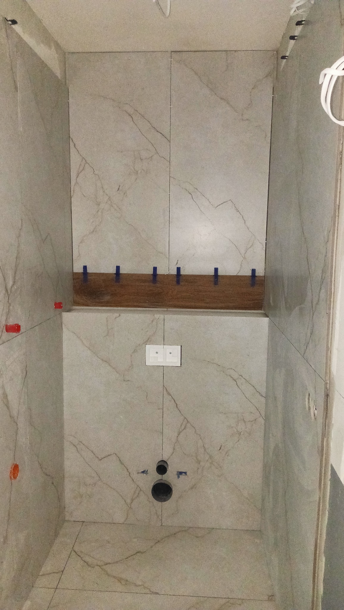 Ściana w trakcie układania płytek imitujących marmur, z widocznymi elementami poziomującymi i otworem na instalację. Etap prac wykończeniowych w łazience.