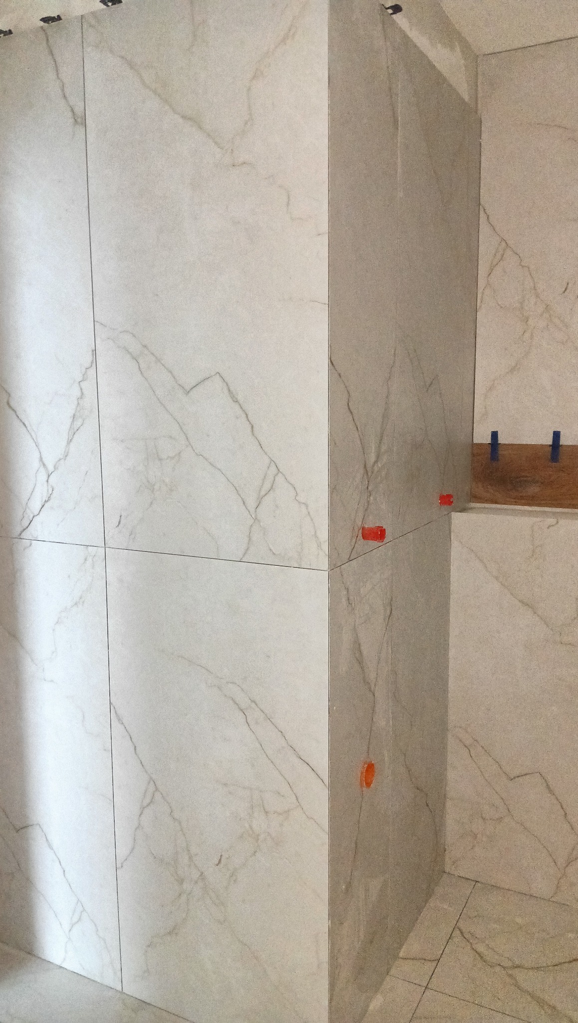 Ściana wyłożona jasnymi płytkami imitującymi marmur, z widocznymi pomarańczowymi i niebieskimi krzyżykami dystansowymi. Widok na narożnik ściany i fragment podłogi.