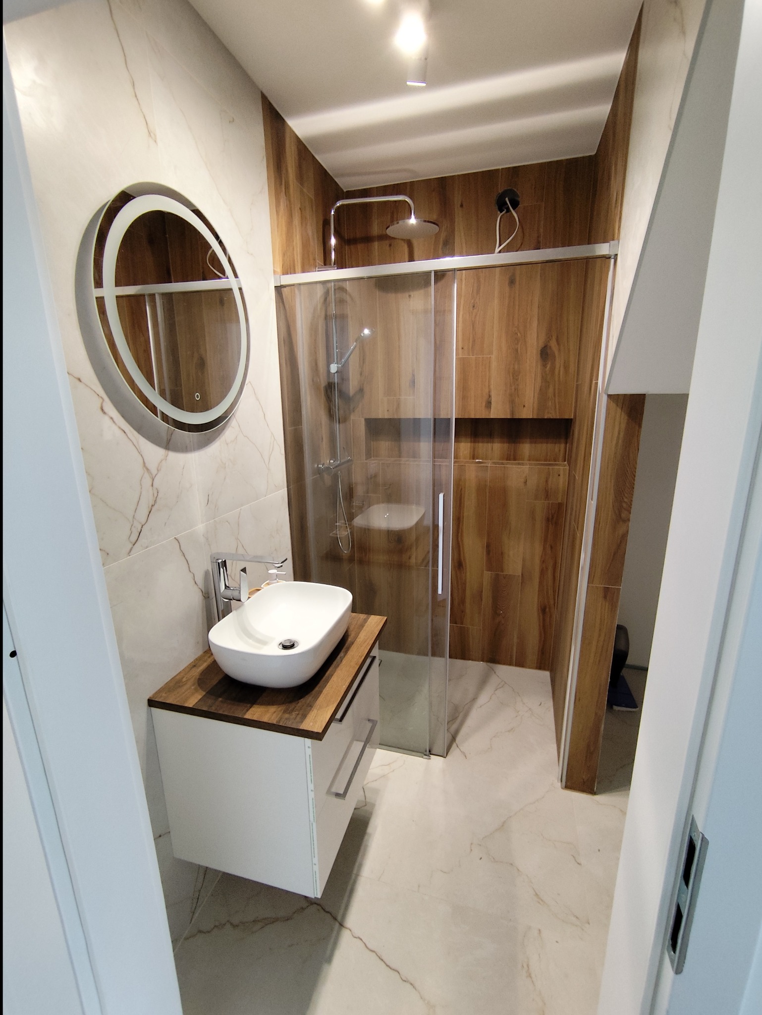 Nowoczesna łazienka z kabiną prysznicową typu walk-in, umywalką nablatową na szafce z drewnianym blatem i okrągłym lustrem z podświetleniem LED.