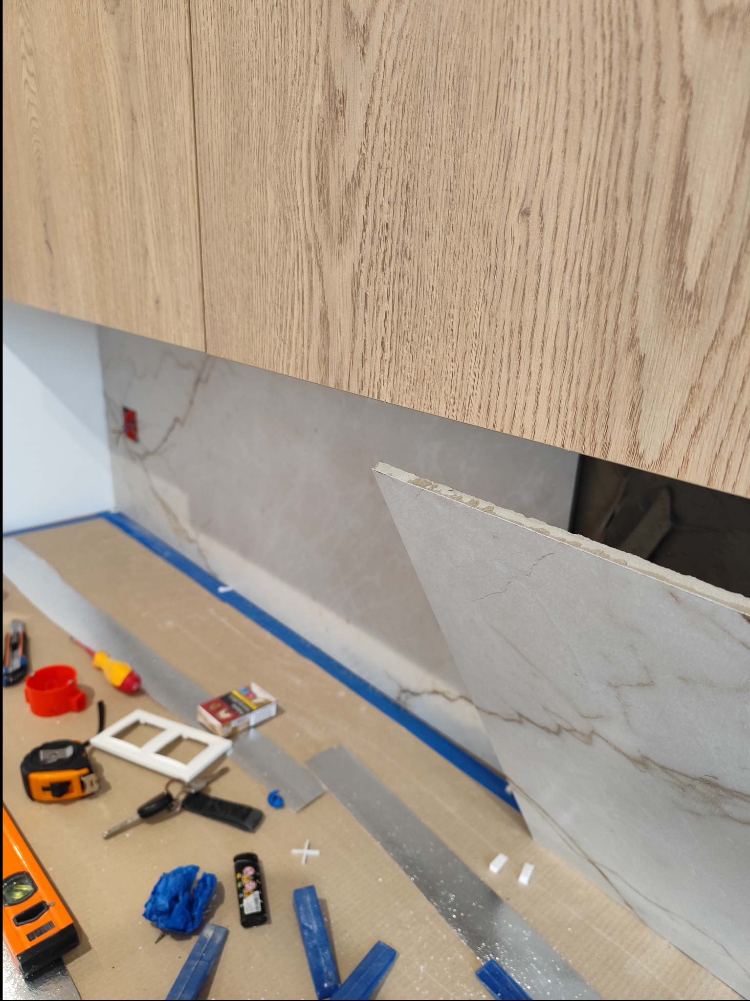 Montaż płytek imitujących marmur na ścianie kuchni pod wiszącymi szafkami. Widoczne narzędzia glazurnicze i fragment nieprzyklejonej płytki.