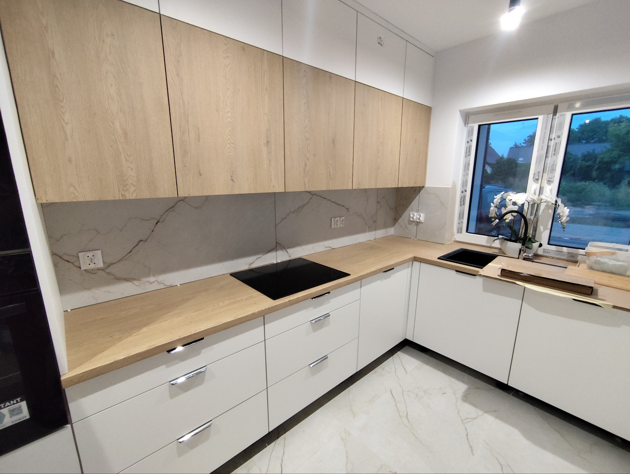 Nowoczesna kuchnia z białymi szafkami i drewnianymi blatami, płyta indukcyjna, marmurowe wykończenie ściany nad blatem, widok na zewnątrz przez okno.