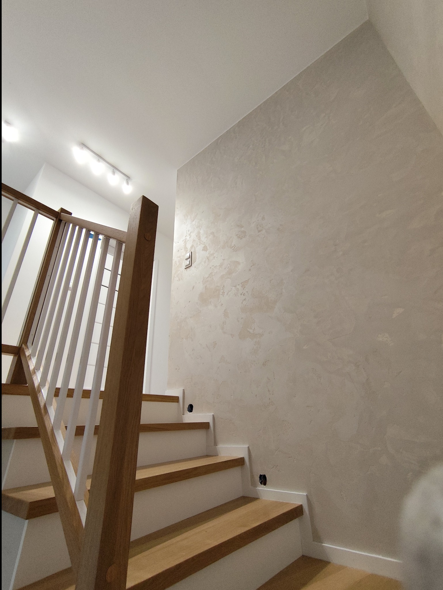Wykończone schody z drewnianymi stopniami i białą balustradą. Ściana w beżowym odcieniu z dekoracyjną fakturą. Widoczne gniazdka elektryczne. Górne oświetlenie.