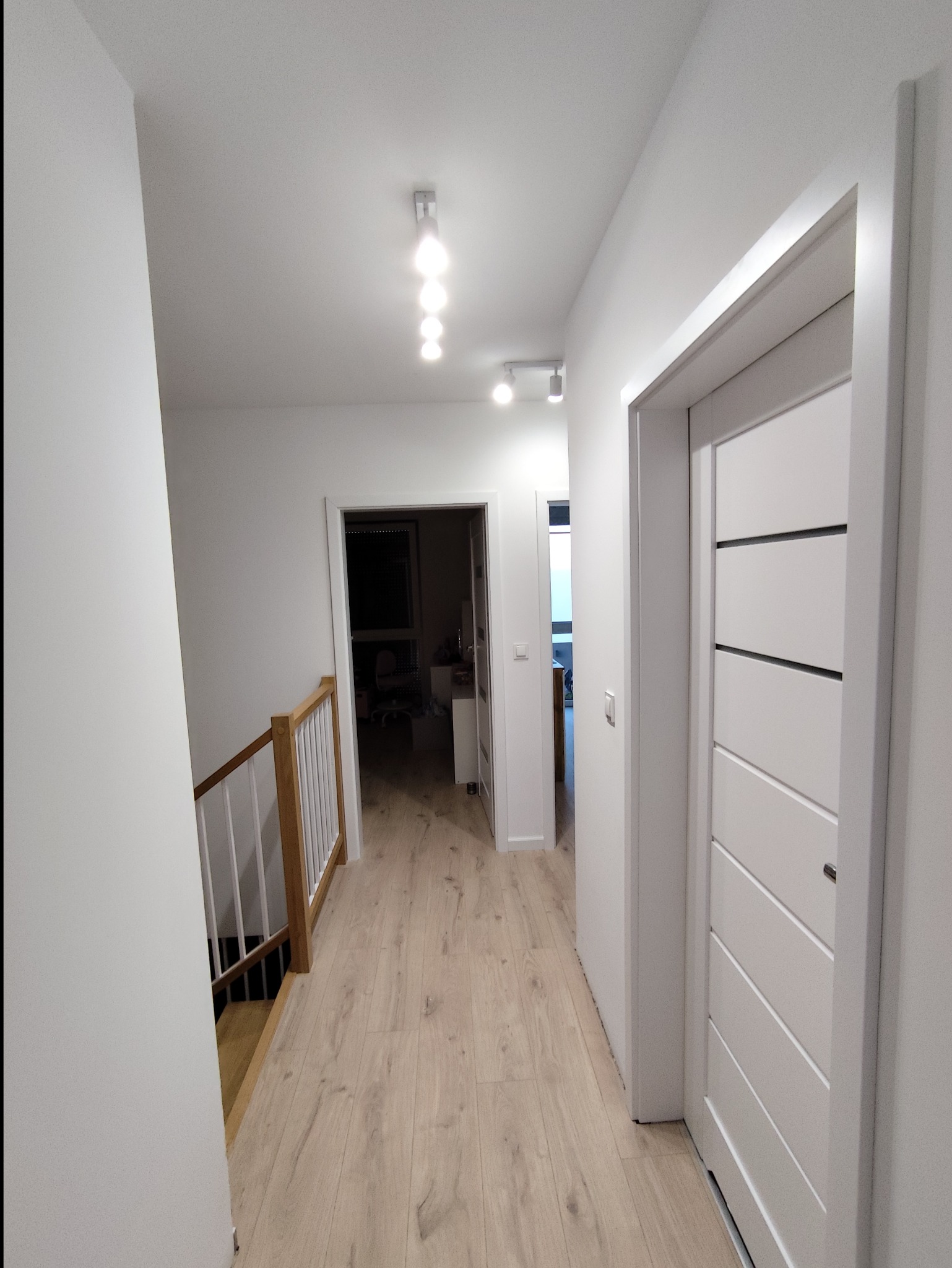 Nowoczesny korytarz z jasnymi panelami, białymi ścianami i drzwiami. Widoczne oświetlenie sufitowe i drewniana balustrada schodów.