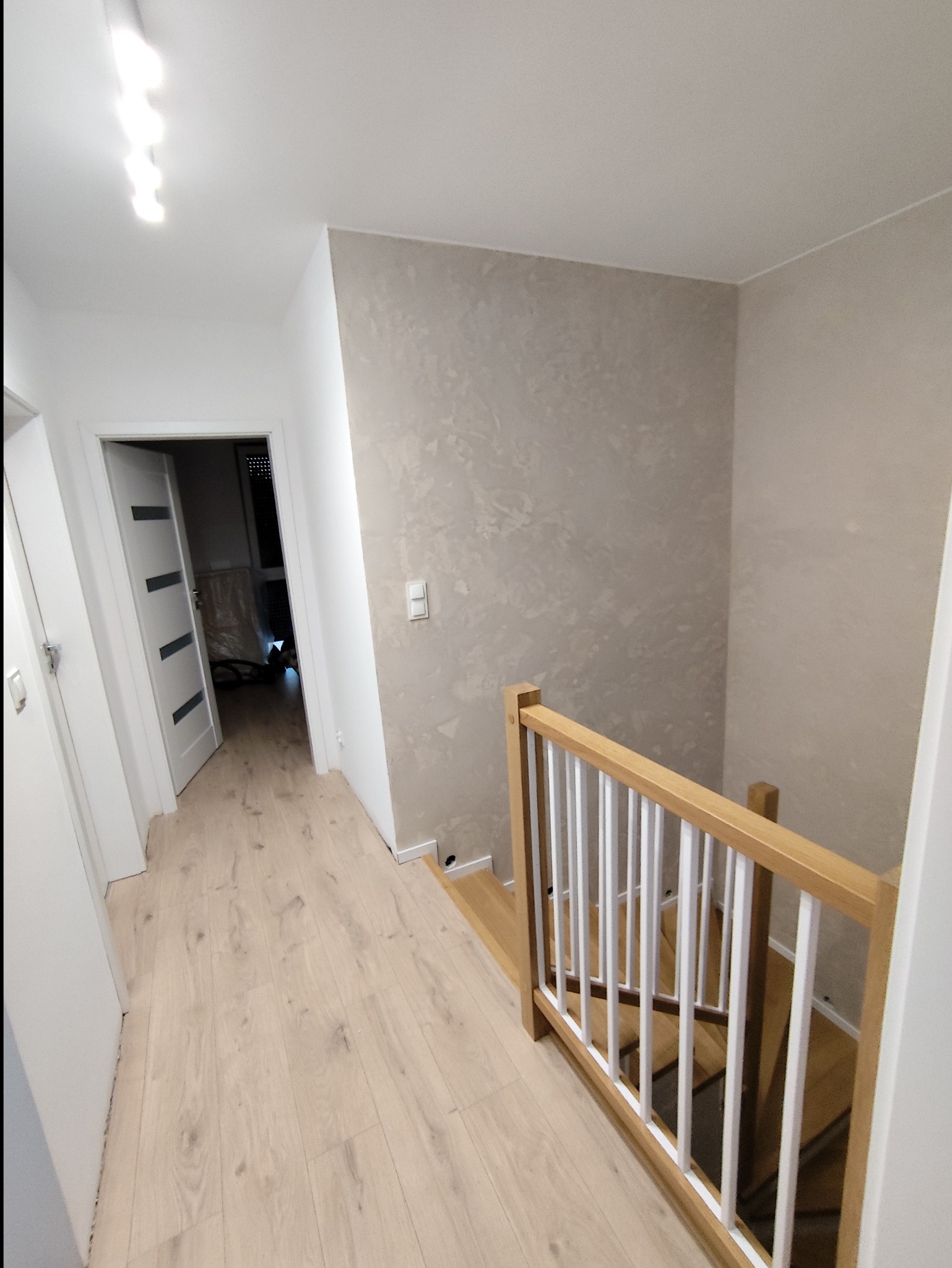 Jasne wnętrze domu: hall z drewnianą podłogą, schody z białą balustradą i kremowa ściana z dekoracyjną fakturą. Widoczne drzwi i oświetlenie sufitowe.
