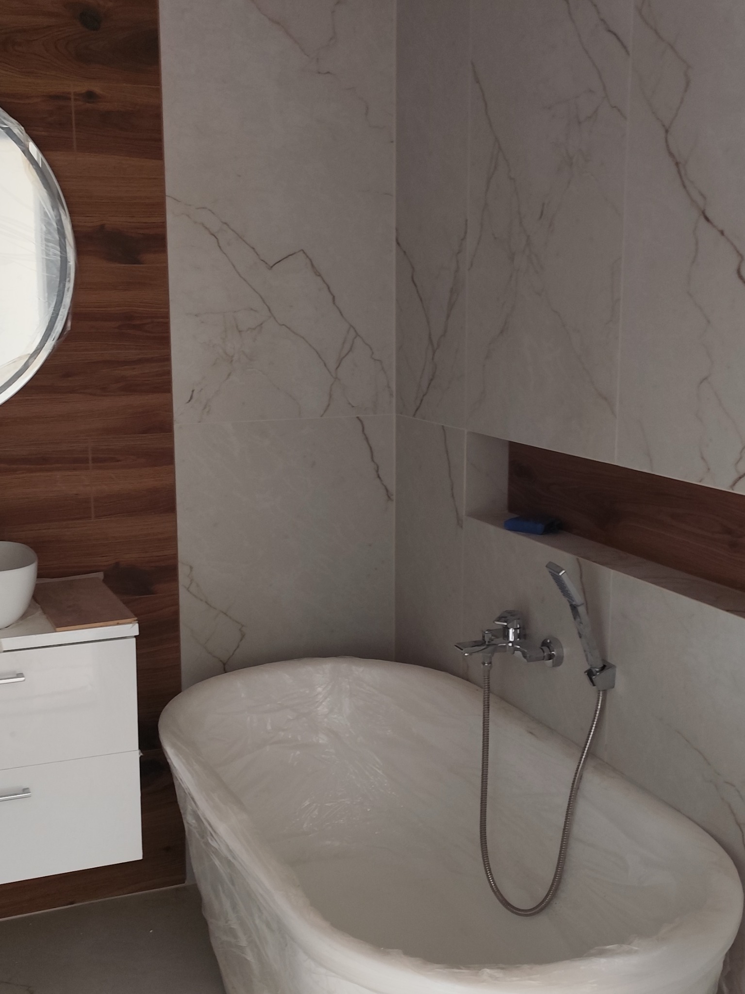 Nowoczesna łazienka w trakcie wykańczania: biała wanna zabezpieczona folią, marmurowe płytki, umywalka z szafką i okrągłe lustro. Widoczny fragment półki w ścianie.