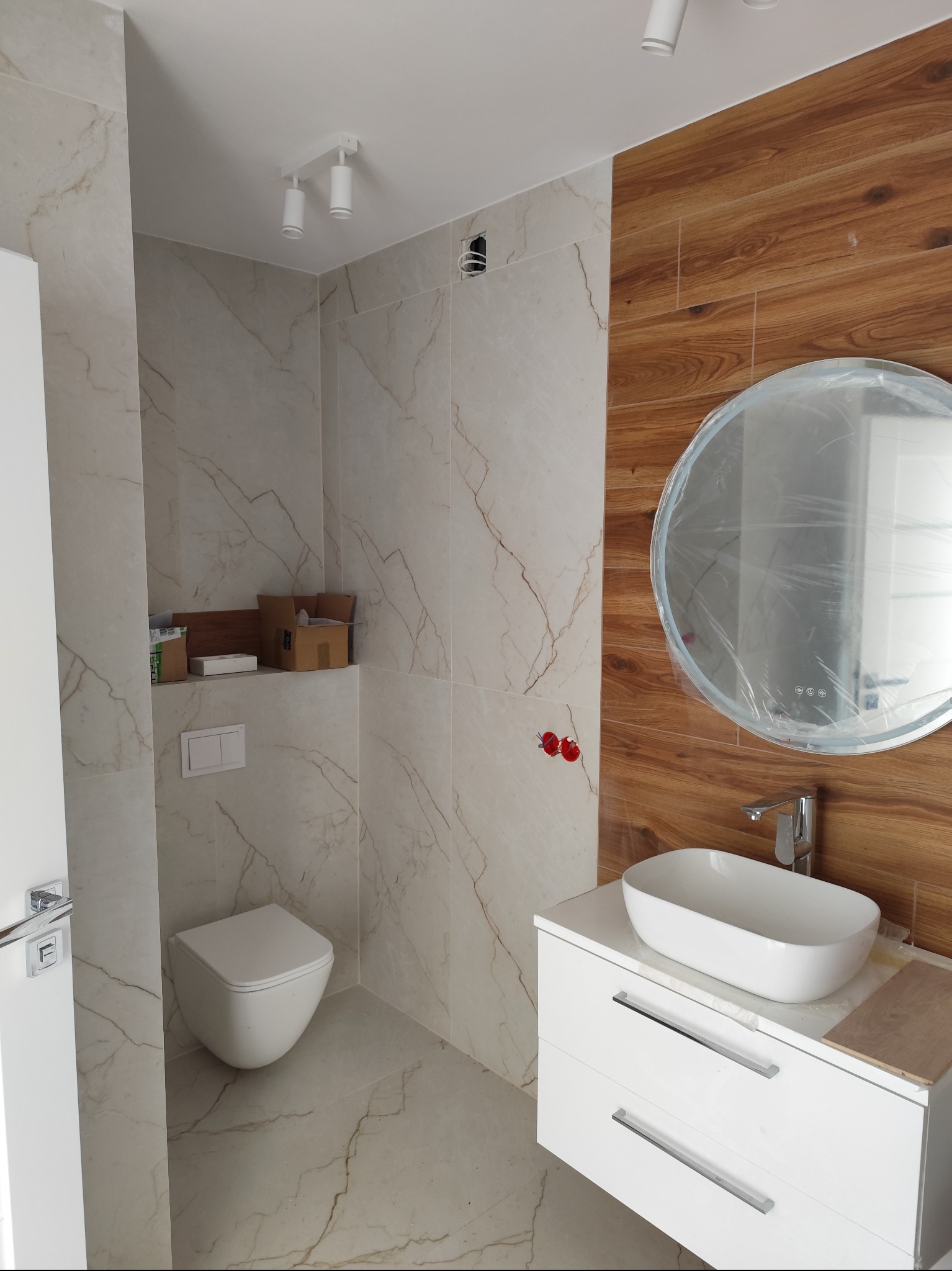 Nowoczesna łazienka z marmurowymi płytkami i drewnianym akcentem. Umywalka nablatowa na białej szafce, okrągłe lustro w folii ochronnej, podwieszana toaleta.
