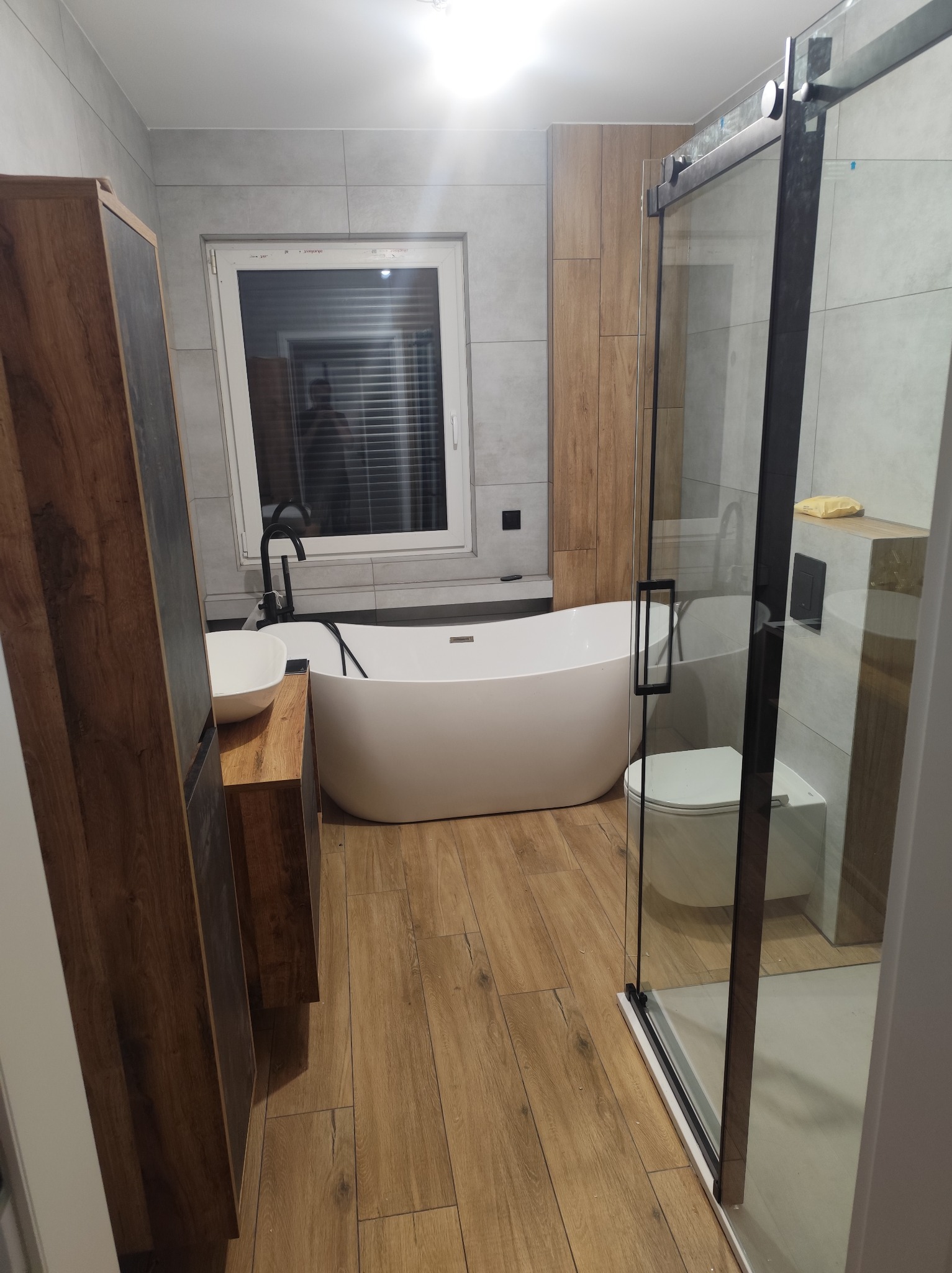 Nowoczesna łazienka z wolnostojącą wanną, kabiną prysznicową ze szklanymi drzwiami i drewnianymi akcentami. Minimalistyczny design, szare płytki i drewniana podłoga.