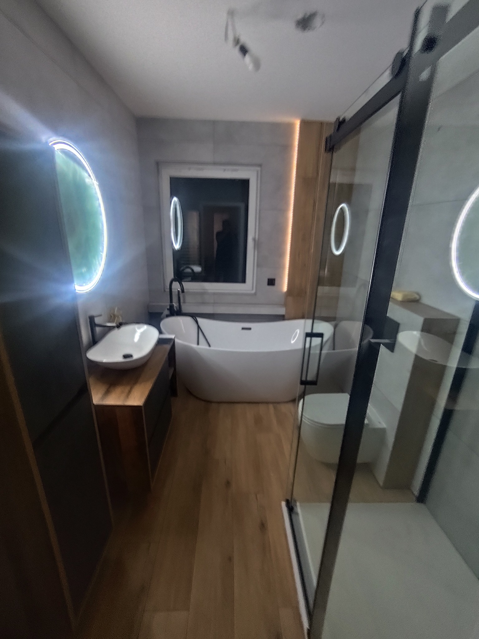 Nowoczesna łazienka z wolnostojącą wanną, umywalką na drewnianym blacie i kabiną prysznicową ze szklanymi drzwiami. Oświetlenie LED podkreśla minimalistyczny design.