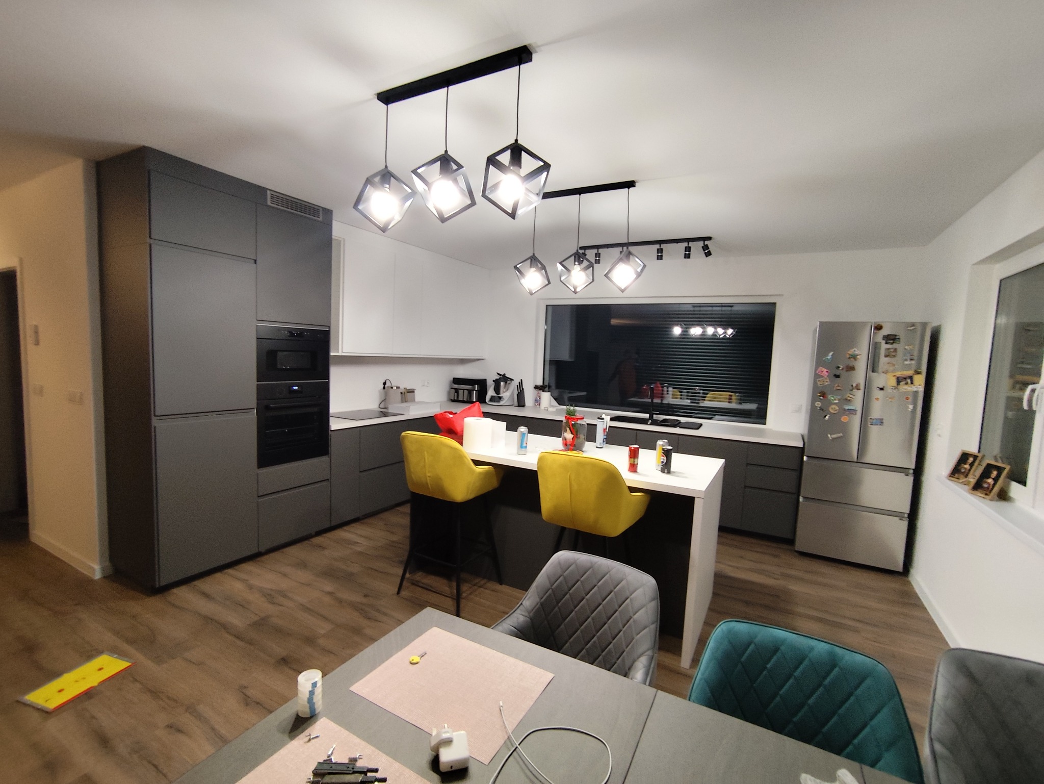 Nowoczesna kuchnia z szarymi szafkami, wyspą z żółtymi krzesłami barowymi i designerskimi lampami. Widoczne drewniane panele podłogowe i srebrna lodówka.