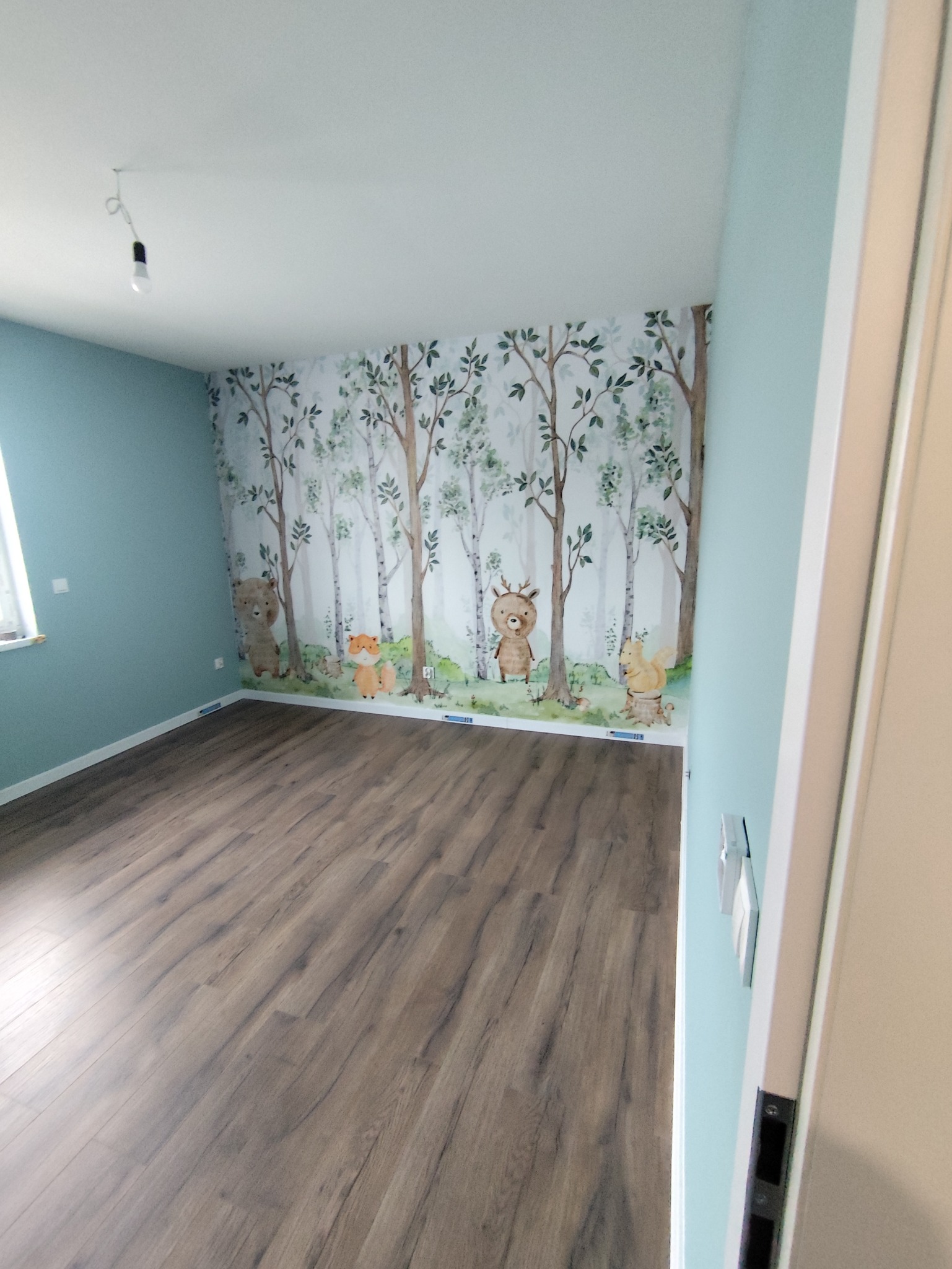 Pokój dziecięcy po remoncie: drewniana podłoga, ściany w kolorze błękitu i tapeta z motywem leśnych zwierząt. Widok na całe pomieszczenie, z perspektywy wejścia.