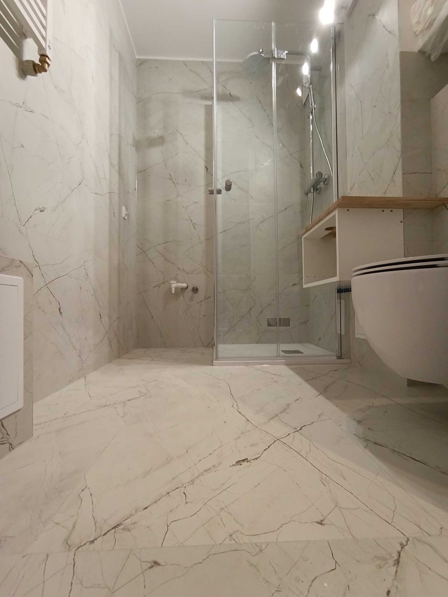 Wykończona łazienka z kabiną prysznicową typu walk-in, białą armaturą i płytkami imitującymi marmur na ścianach i podłodze. Minimalistyczny design.