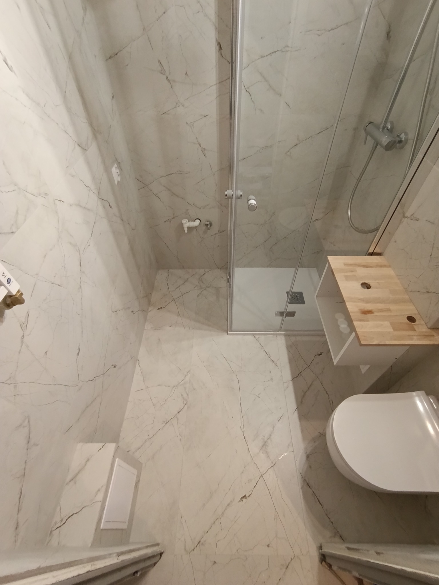 Wykończona łazienka z kabiną prysznicową, toaletą i marmurowymi płytkami. Uwagę zwraca drewniany blat w kabinie.