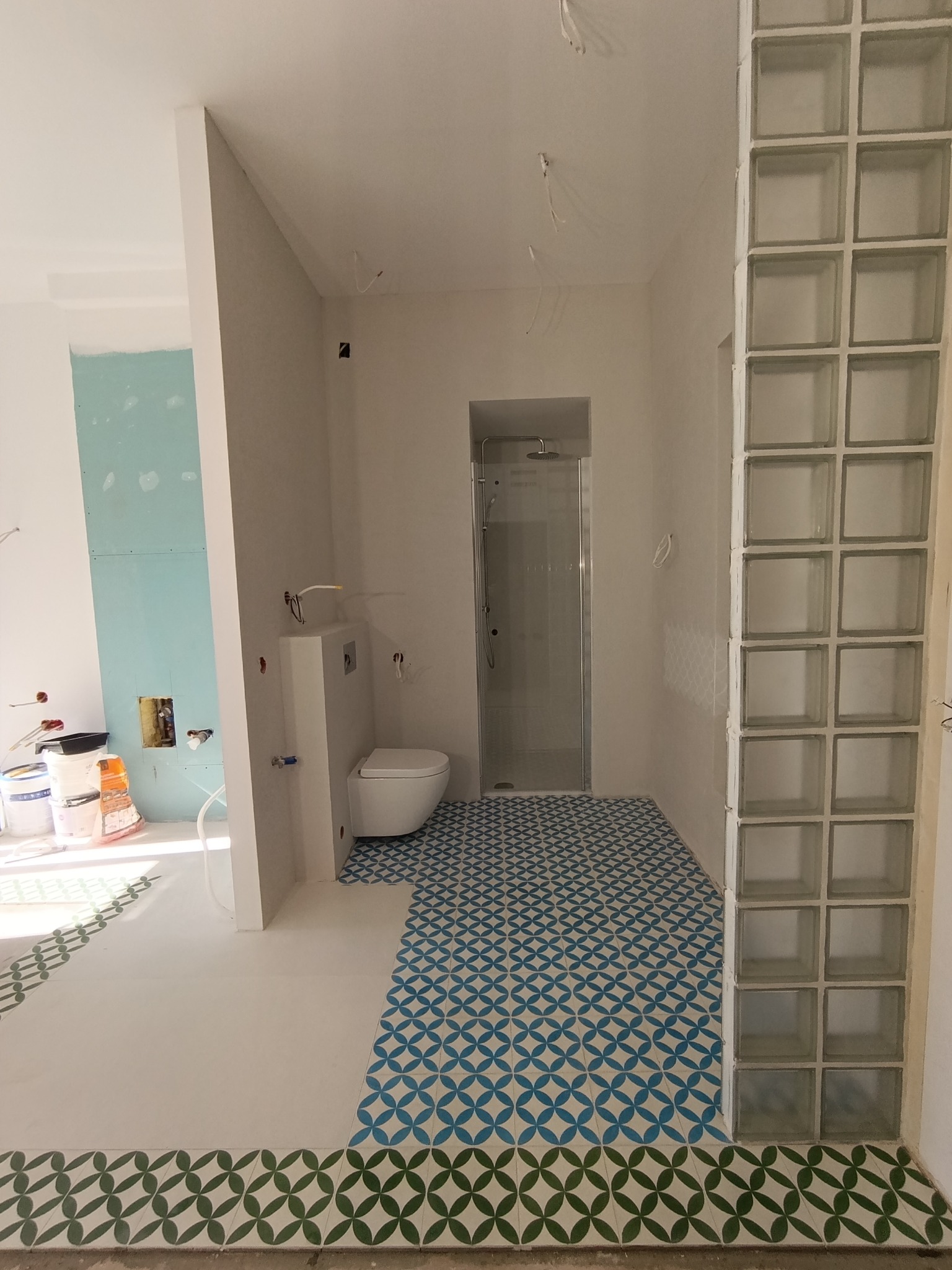 Wnętrze łazienki w trakcie remontu: ułożone, wzorzyste płytki podłogowe, kabina prysznicowa, toaleta podwieszana, ściana z luksferów. Widoczne niedokończone prace wykończeniowe.