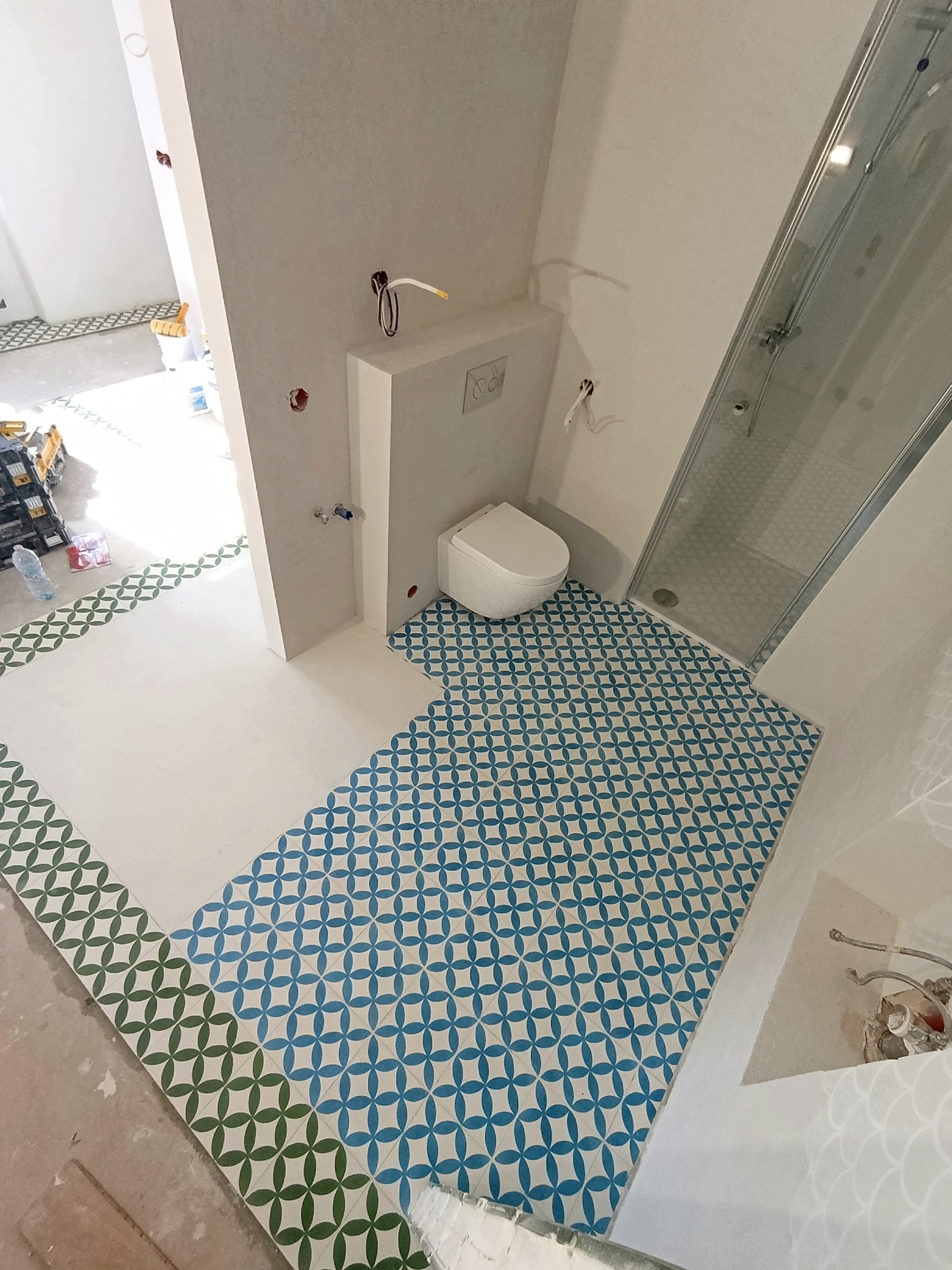 Wnętrze łazienki w trakcie prac wykończeniowych. Ułożona, wzorzysta podłoga w odcieniach błękitu i zieleni. Widoczna kabina prysznicowa i toaleta w zabudowie.
