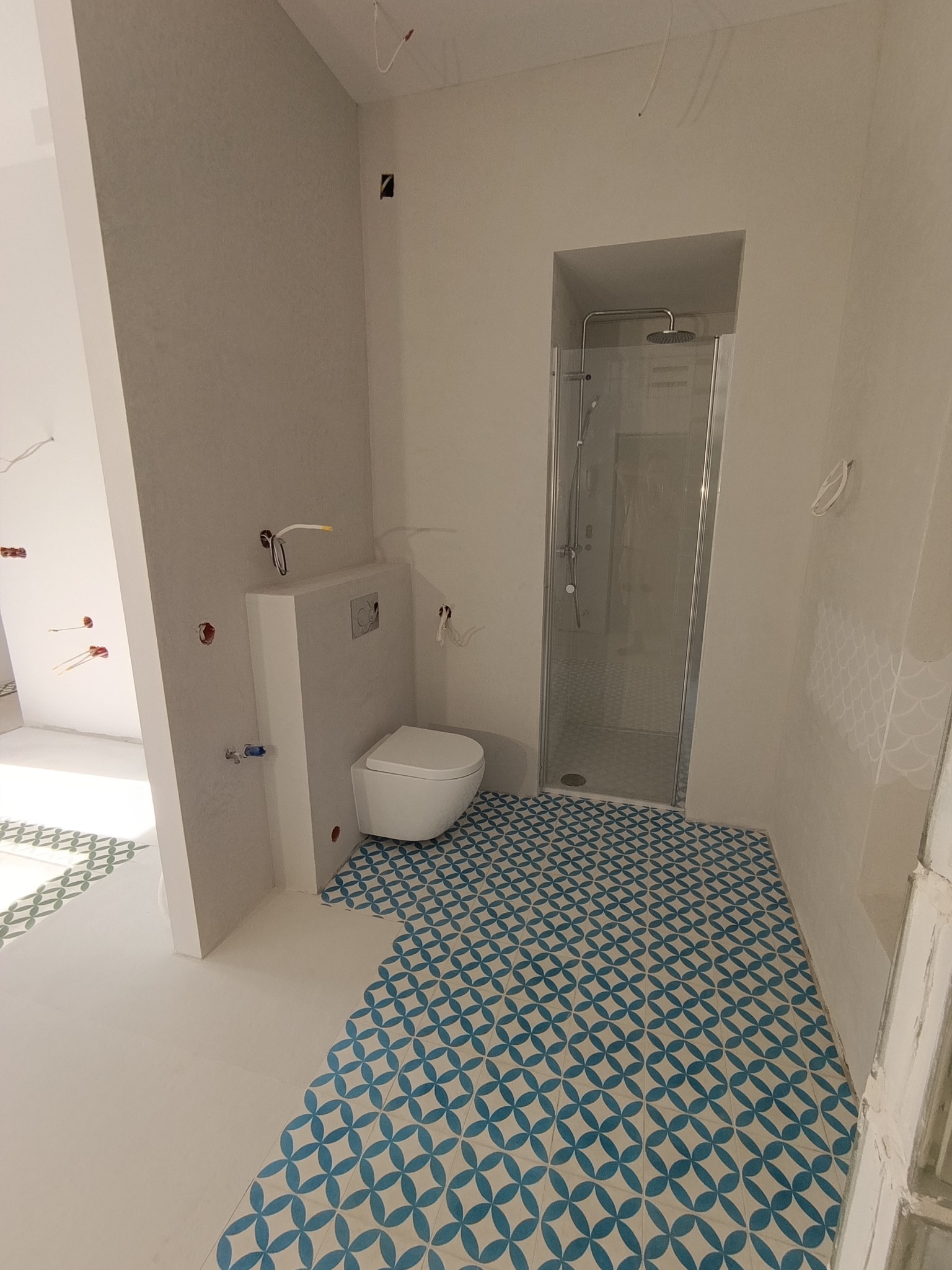 Biała łazienka z kabiną prysznicową i toaletą w zabudowie. Podłoga wyłożona niebiesko-białymi płytkami w geometryczny wzór. Widoczne instalacje przed wykończeniem.