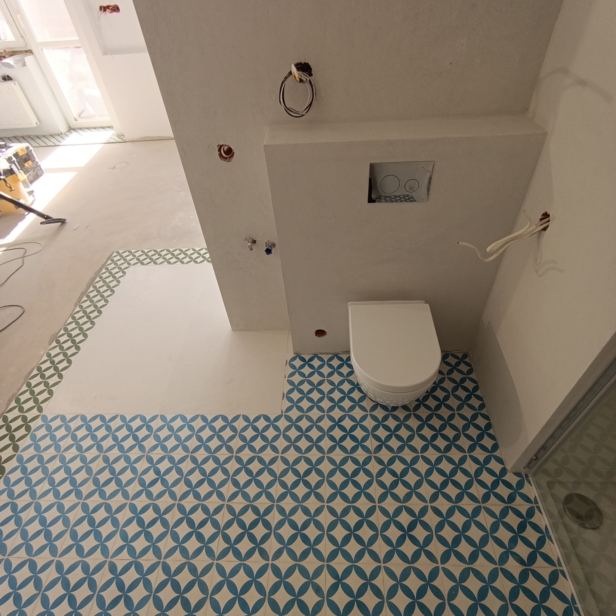 Wnętrze łazienki w trakcie wykańczania: ułożone płytki podłogowe z geometrycznym wzorem, częściowo wykończona ściana z podtynkową toaletą, widoczne instalacje.