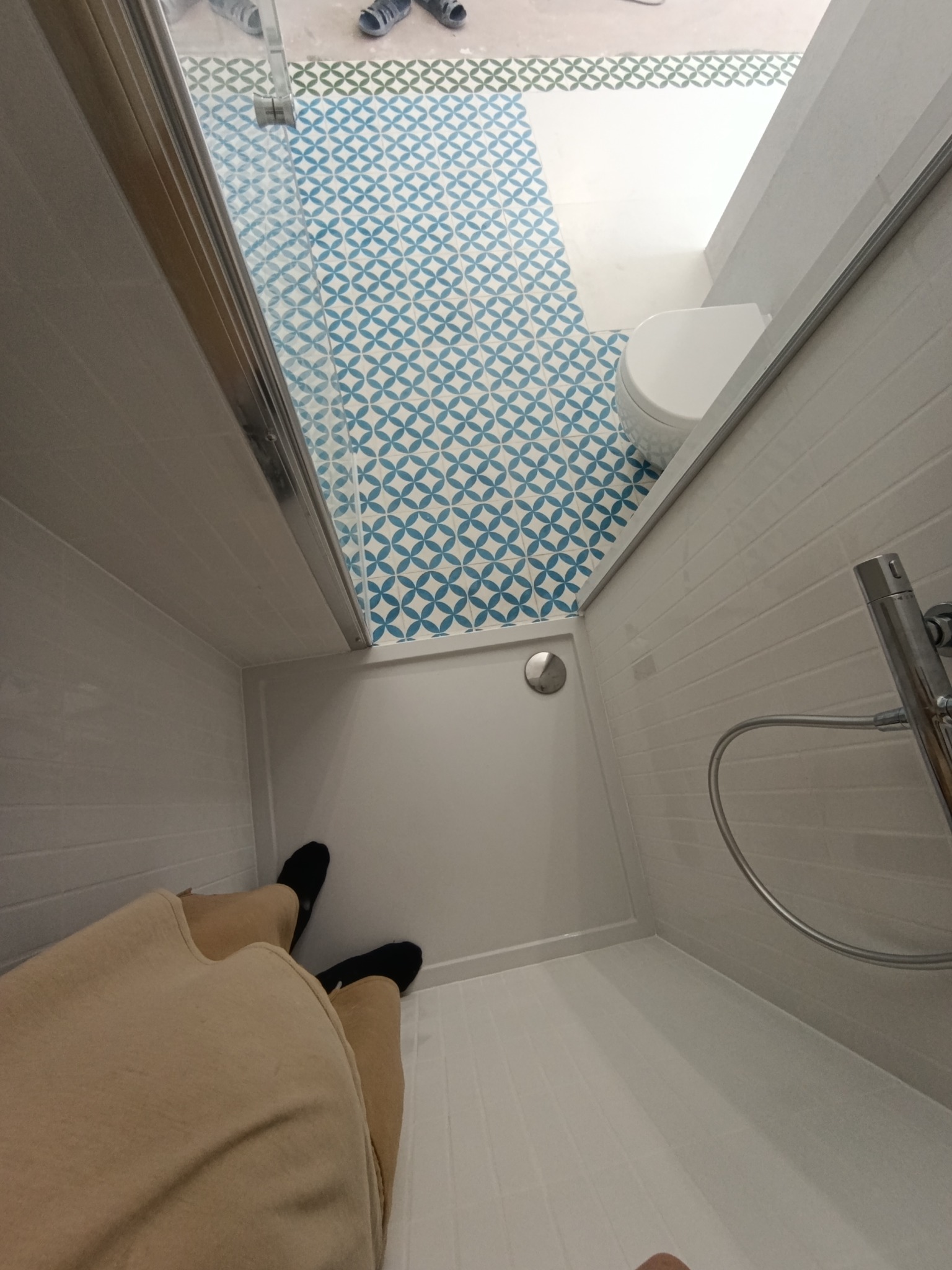 Nowoczesna łazienka z kabiną prysznicową i wzorzystą, niebiesko-białą podłogą. Widok z góry na kabinę, fragment toalety i nogi osoby w czarnych skarpetkach.