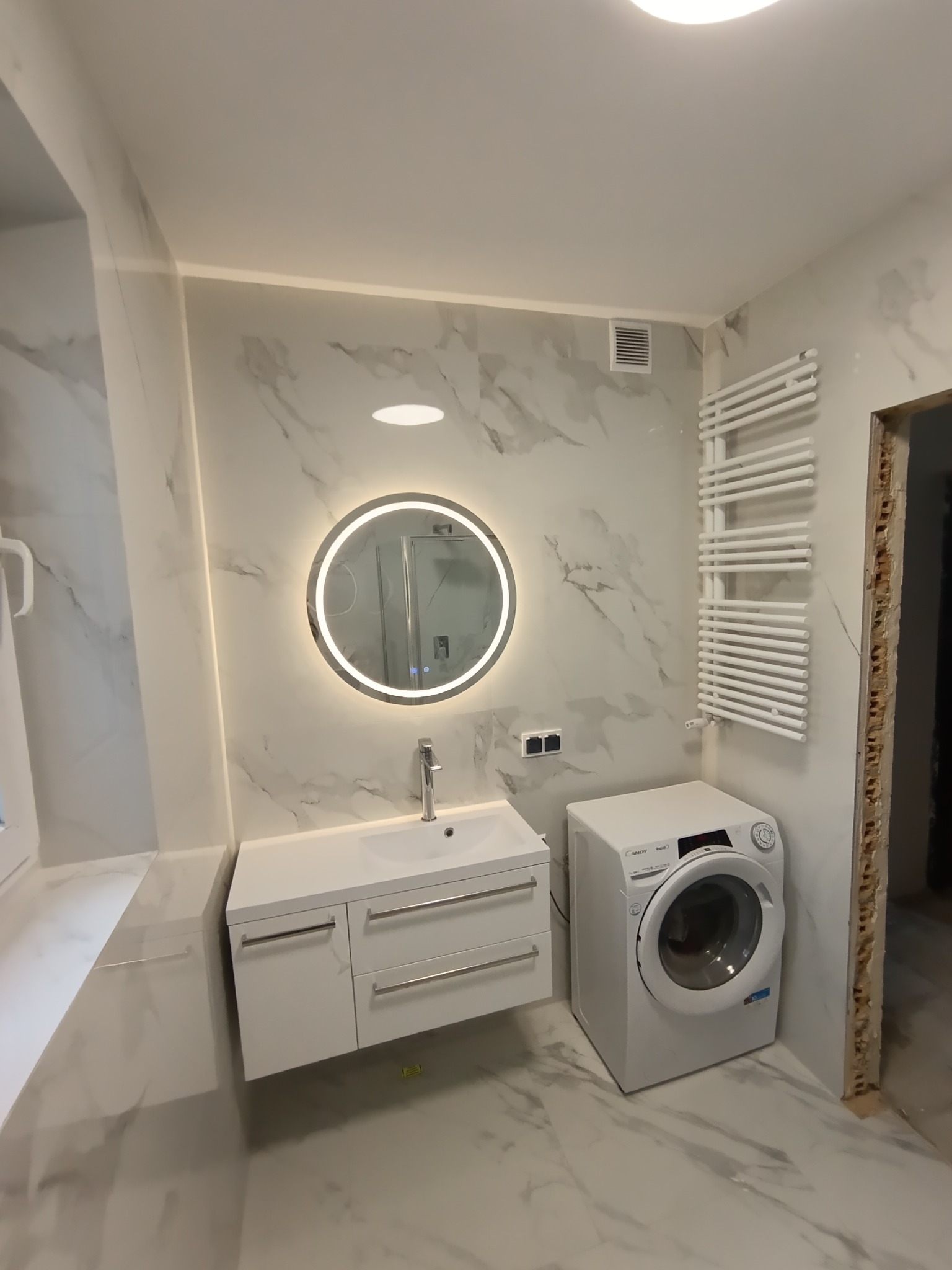 Wykończona łazienka w bieli: umywalka z szafką, okrągłe lustro z podświetleniem LED, grzejnik drabinkowy i pralka. Płytki imitujące marmur na ścianach i podłodze.
