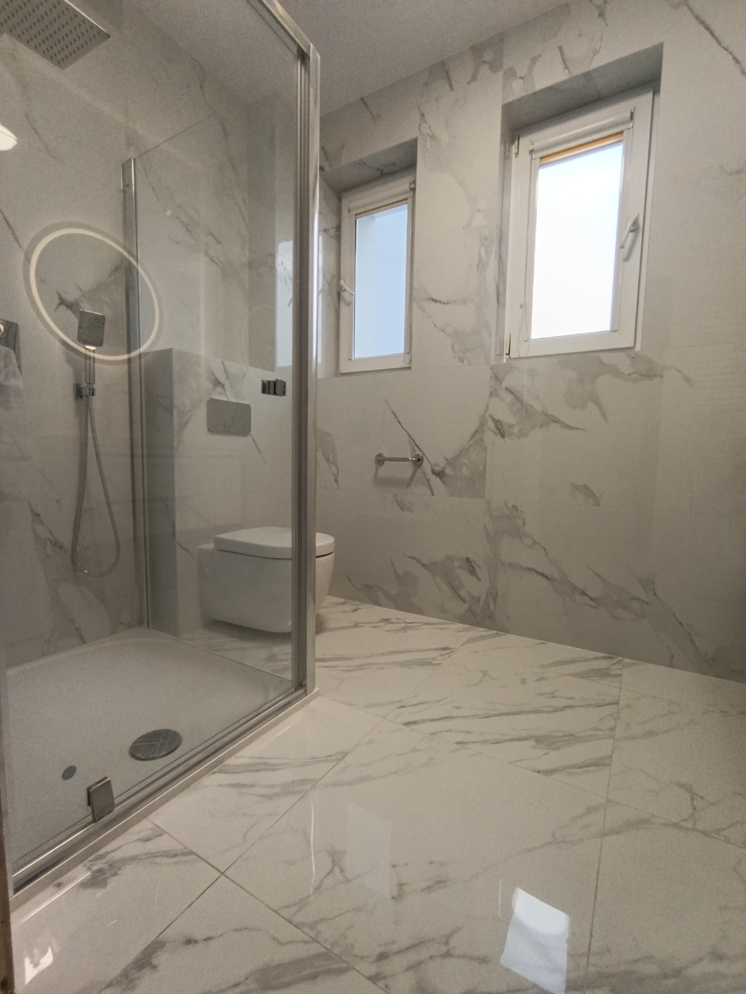 Nowoczesna łazienka z kabiną prysznicową typu walk-in, wykończona białymi płytkami imitującymi marmur. Dwa okna wpuszczają naturalne światło.