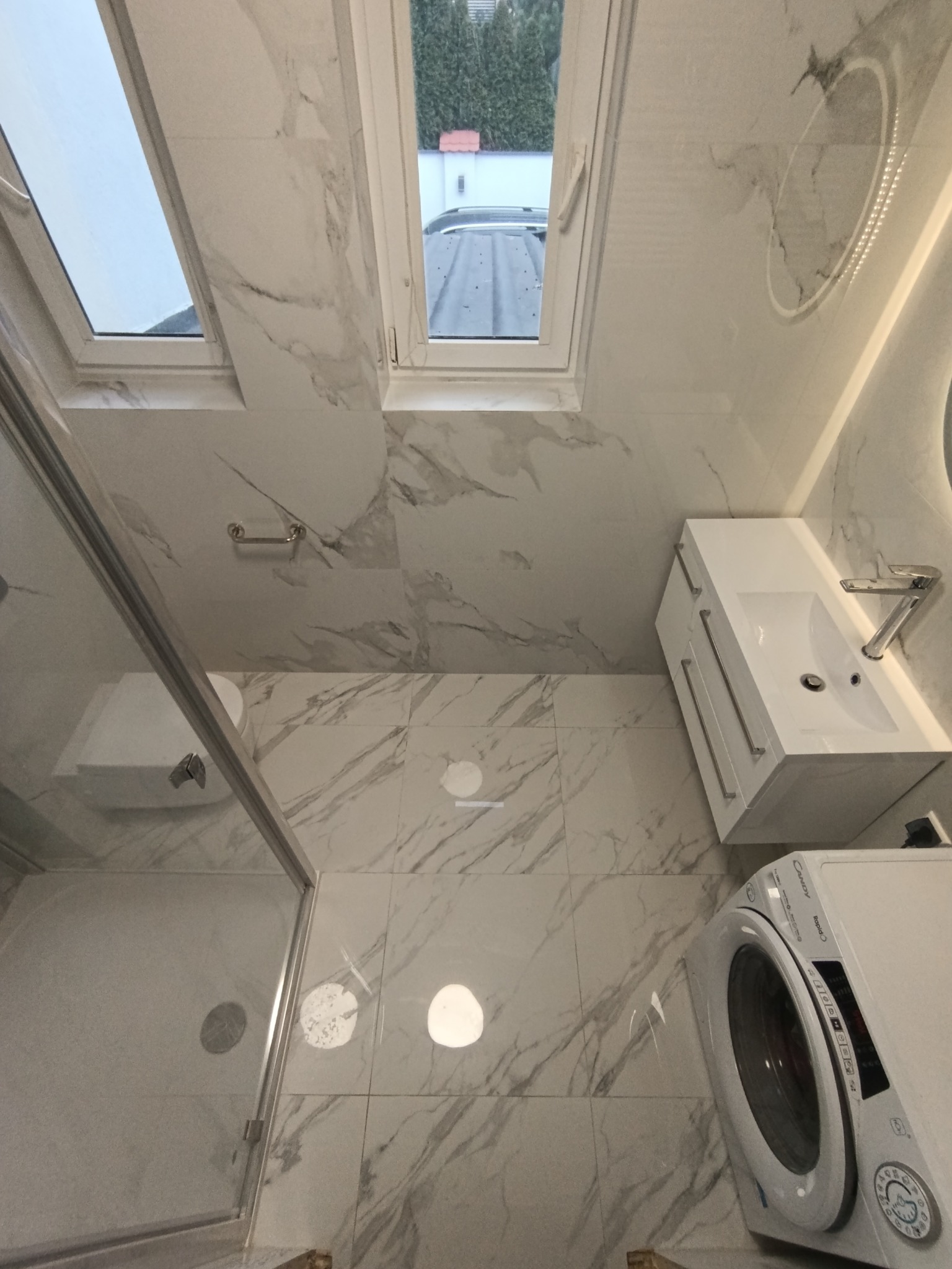 Jasna łazienka z marmurowymi płytkami, kabiną prysznicową, umywalką z szafką i pralką. Widok z góry podkreśla przestronność i nowoczesny design.