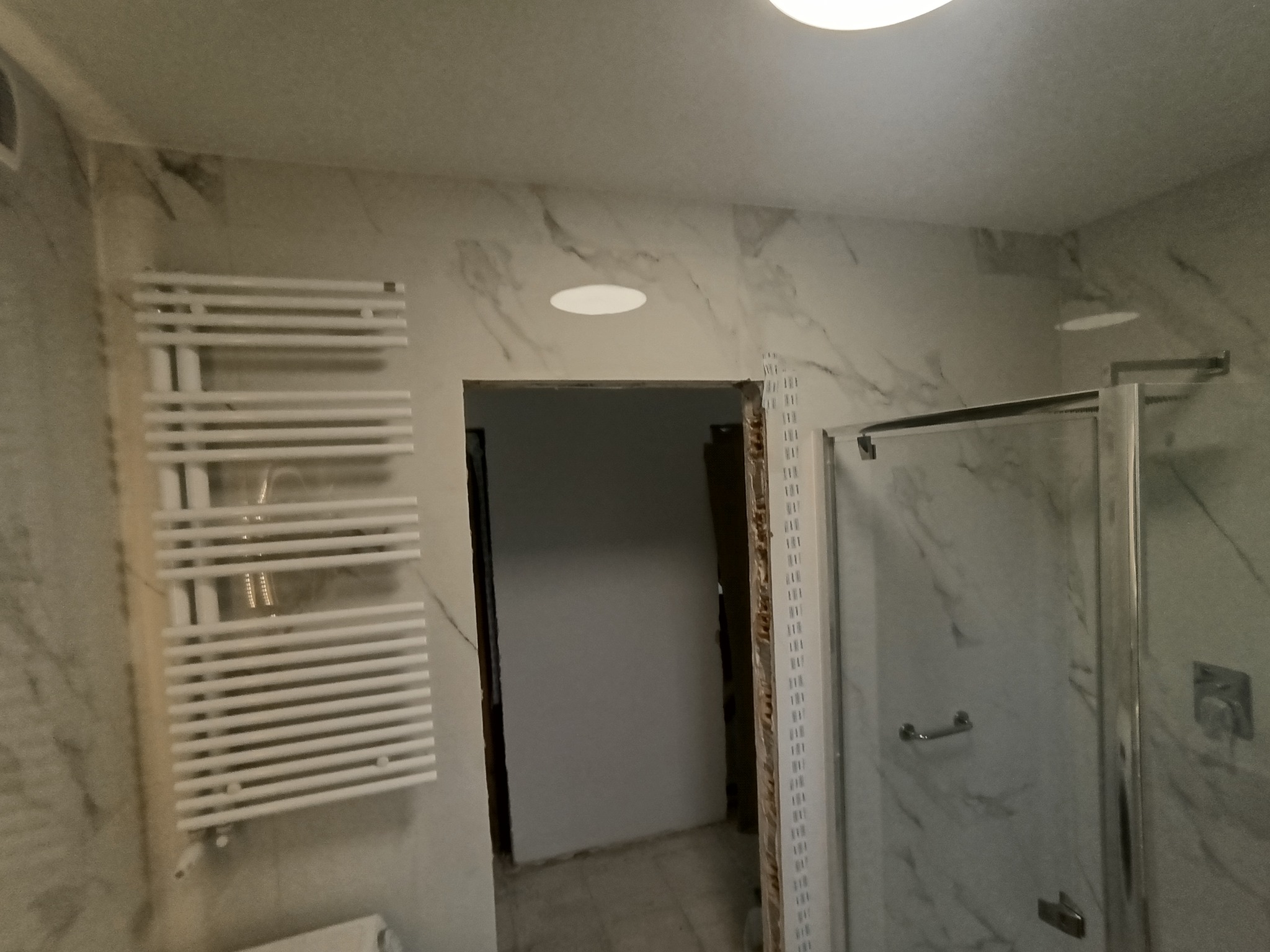 Biała łazienka w trakcie wykończenia: marmurowe płytki, grzejnik drabinkowy i kabina prysznicowa z widocznym wejściem do innego pomieszczenia.