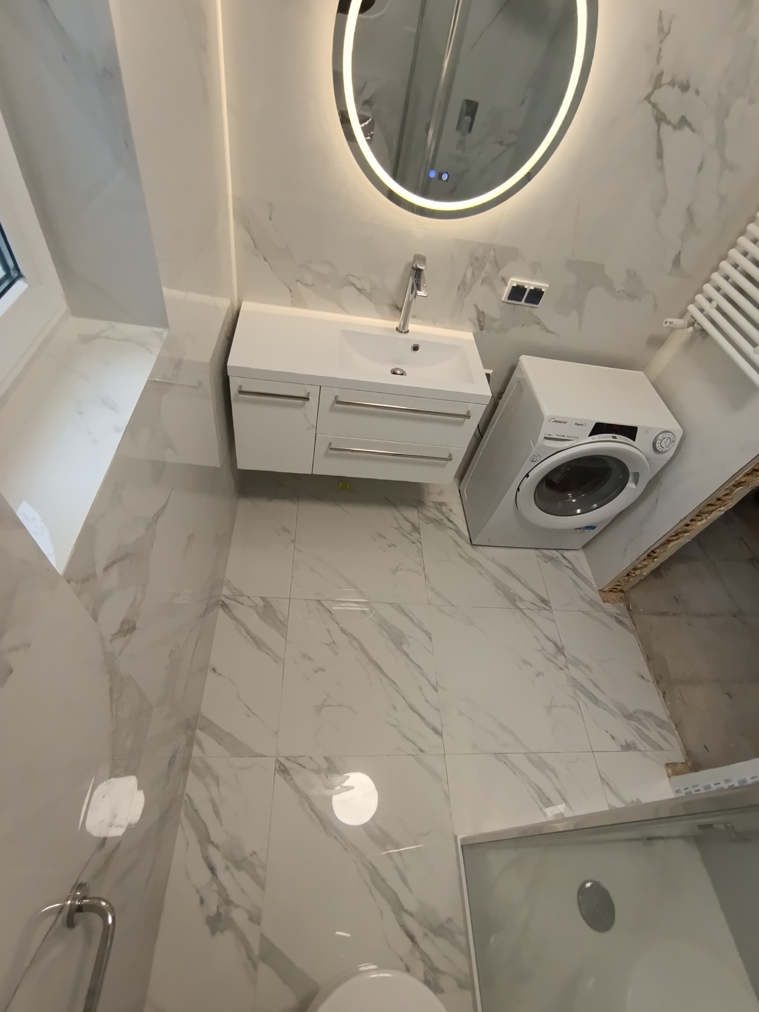 Nowoczesna łazienka w Warszawie z marmurowymi płytkami, okrągłym lustrem z podświetleniem LED, białą szafką z umywalką i pralką. Minimalistyczny design.