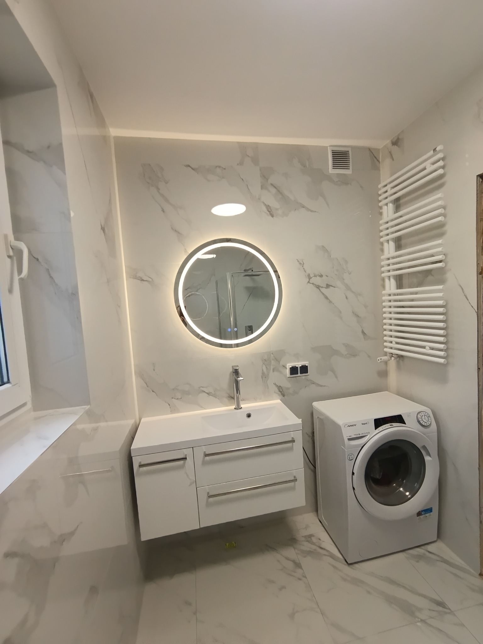 Nowoczesna łazienka w bieli: marmurowe płytki, okrągłe lustro z podświetleniem LED, biała szafka pod umywalką i pralka. Minimalistyczny grzejnik drabinkowy.