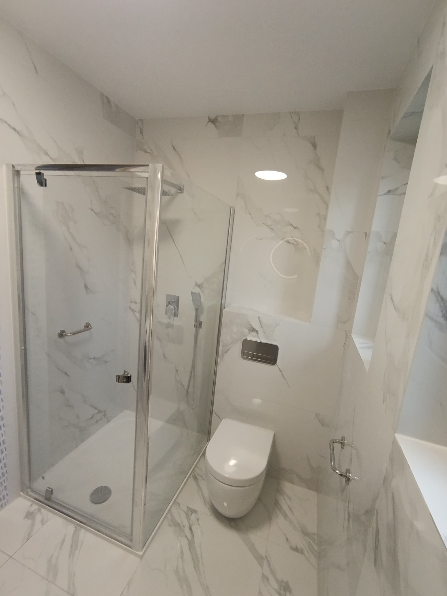Nowoczesna łazienka w bieli i szarości z kabiną prysznicową ze srebrnymi okuciami, wiszącą toaletą i marmurowymi płytkami na ścianach i podłodze.