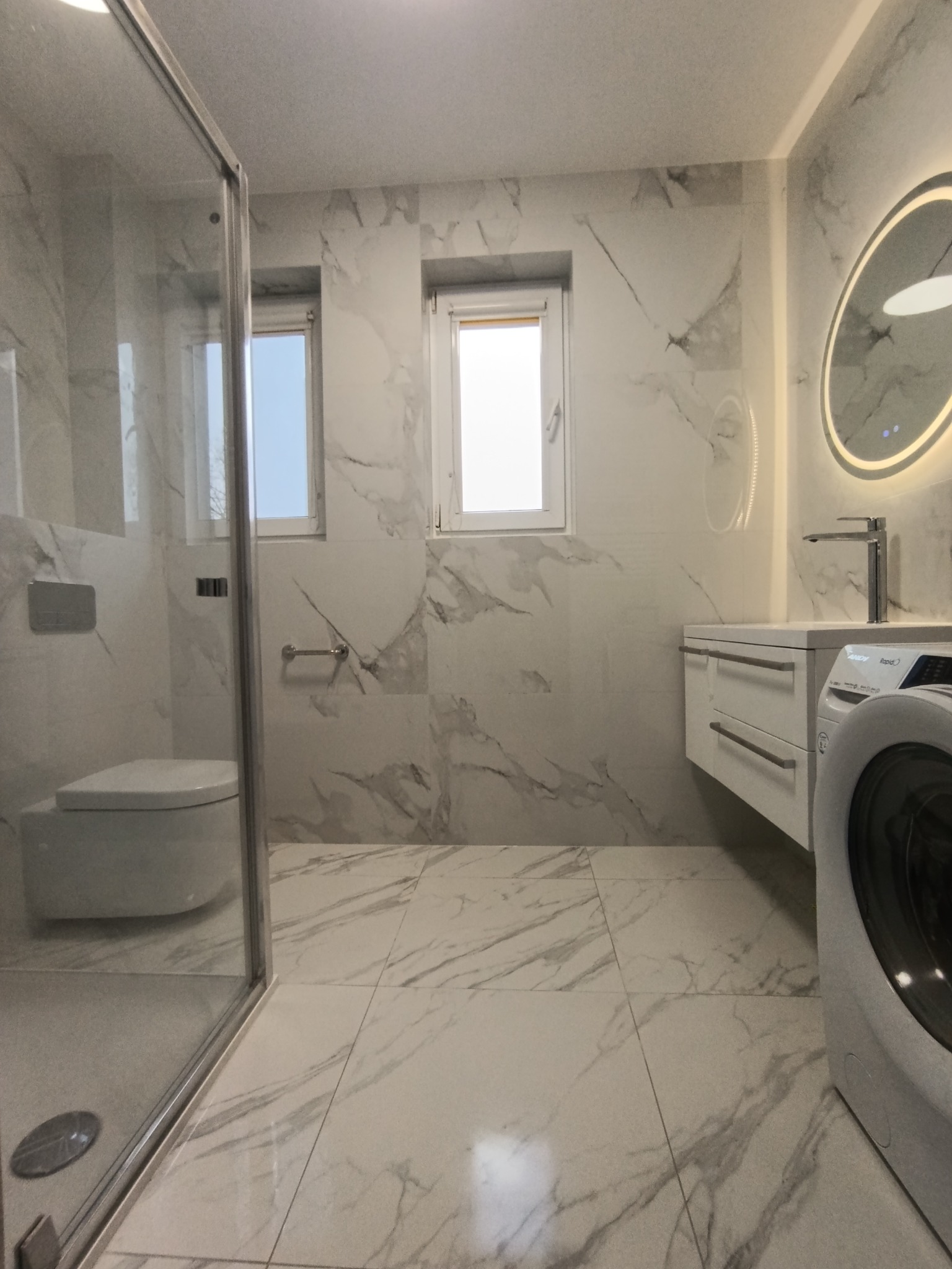 Nowoczesna łazienka z kabiną prysznicową typu walk-in, umywalką z szafką, podświetlanym lustrem i pralką. Białe marmurowe płytki na ścianach i podłodze.