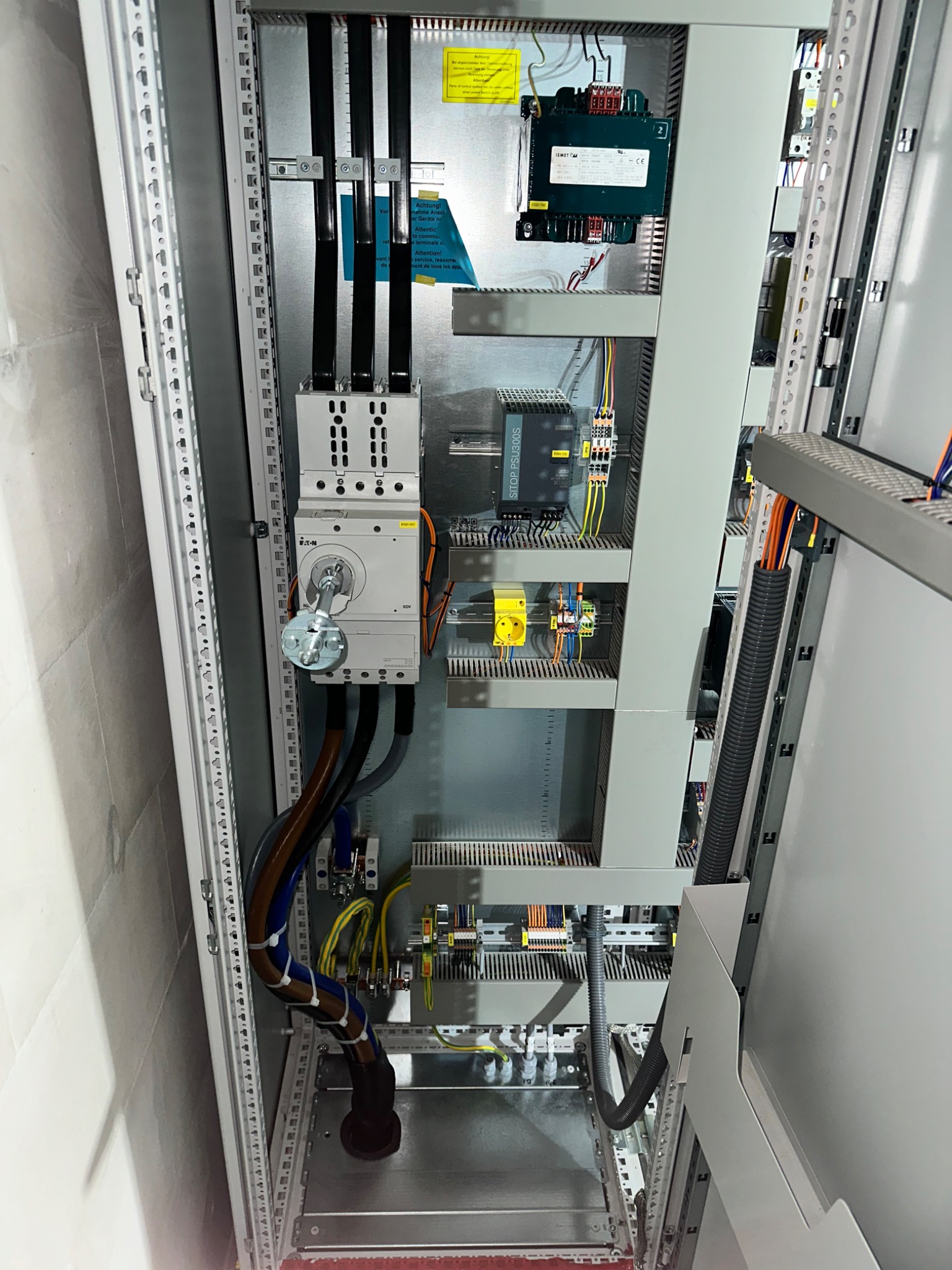 Szara szafa elektryczna z widocznymi kablami, bezpiecznikami i innym osprzętem. Widoczne profesjonalne podłączenia.