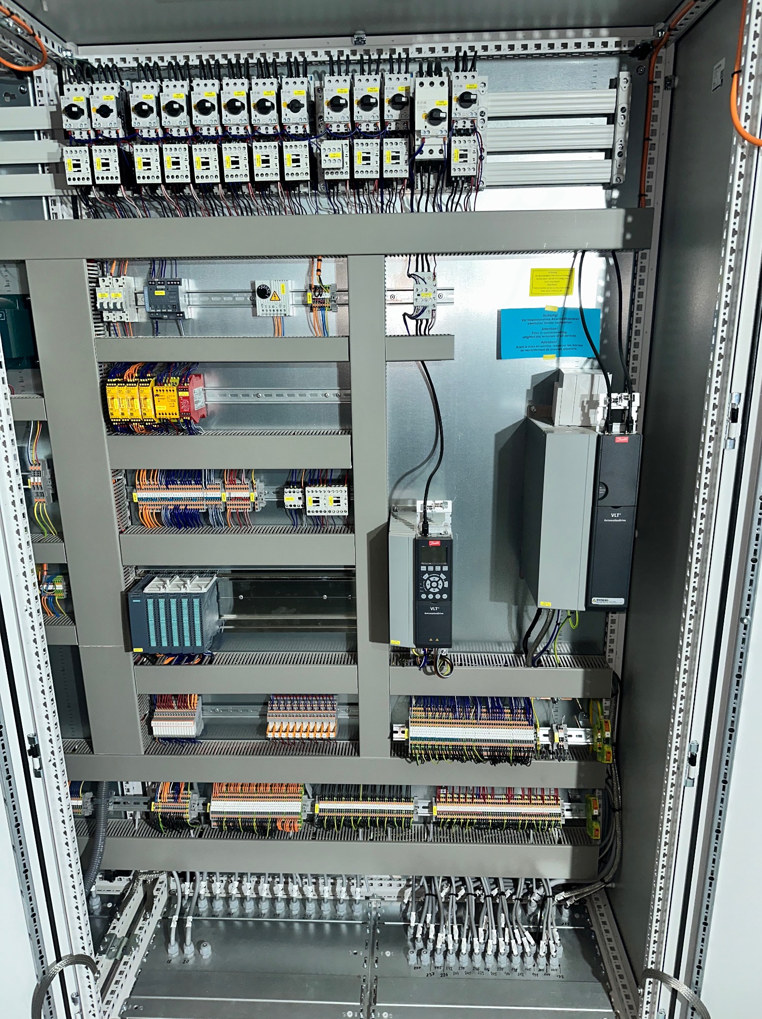 Rozdzielnica elektryczna z licznymi bezpiecznikami, sterownikami PLC, modułami i okablowaniem. Widok na wnętrze szafy sterowniczej z szyną DIN i przewodami.