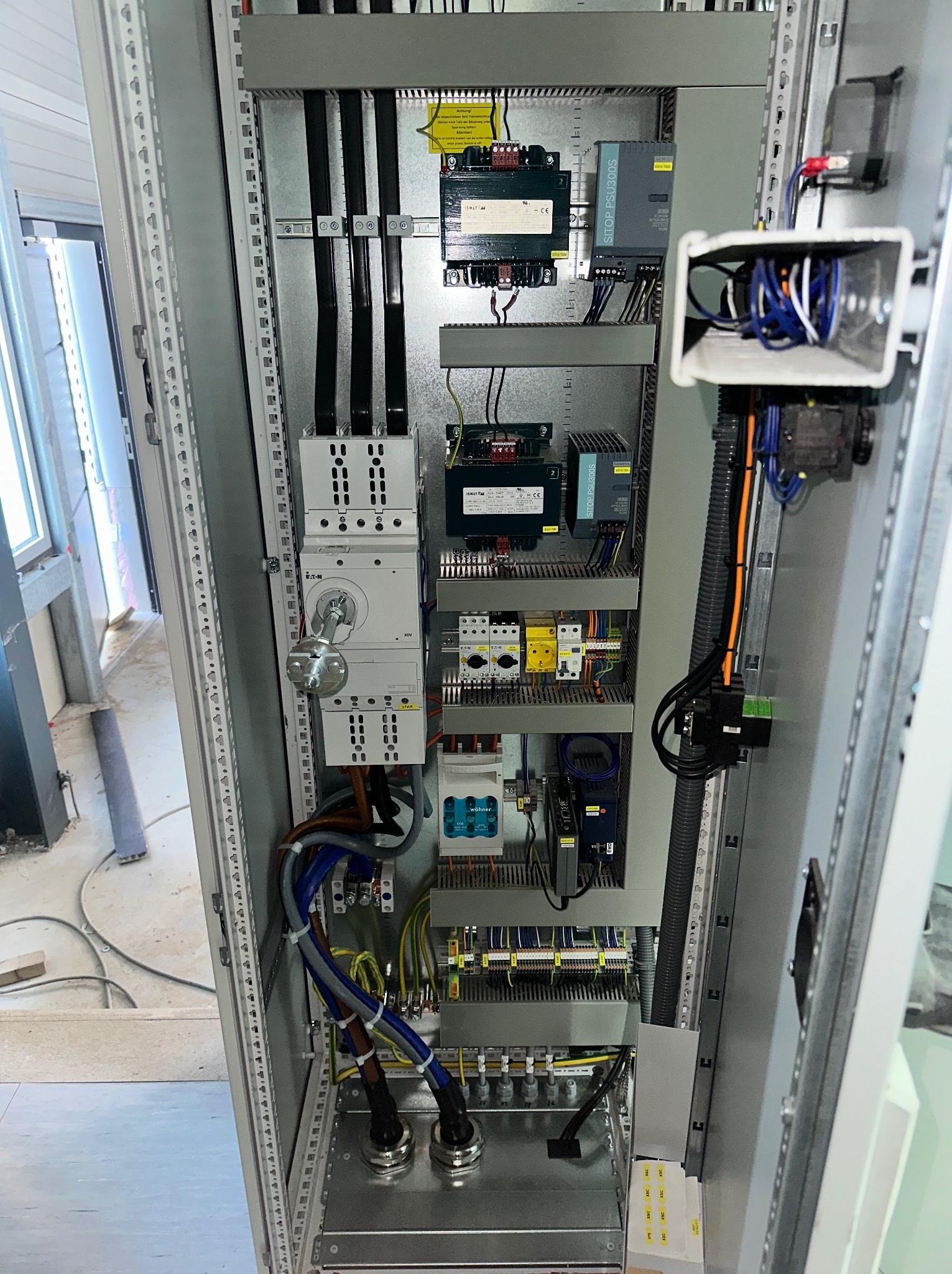 Rozdzielnica elektryczna w metalowej obudowie z widocznymi przewodami, bezpiecznikami i innymi elementami instalacji. Widoczne komponenty Eaton i Siemens.